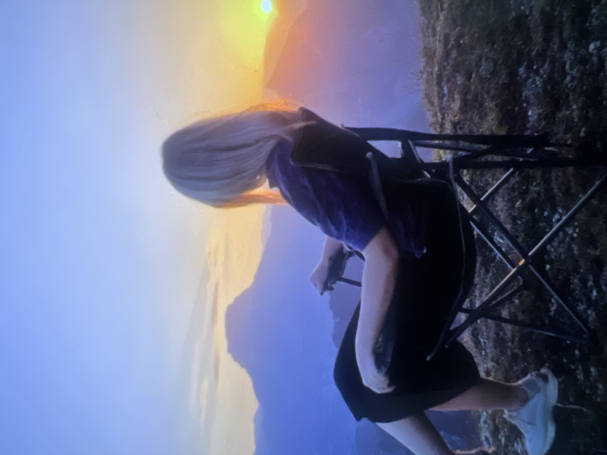 The image displays a panoramic view of a mountainous landscape during either sunrise or sunset, with a person seated on a folding chair in the foreground. The person, seen from behind and slightly to the side, has long blonde hair and is wearing a dark top and dark bottoms. They are seated on a dark metal folding chair, seemingly observing the vista.
The sky is a vibrant gradient, transitioning from a brilliant yellow and orange hue on the left, where the sun is low on the horizon, to softer blues and purples across the rest of the sky. Silhouetted mountains, appearing in shades of dark blue and purple, dominate the mid-ground, extending across the frame. The foreground on the right shows textured, dark ground, possibly sparse vegetation on a rocky outcrop, suggesting an elevated viewpoint.
The scene conveys a sense of tranquility and appreciation for nature, capturing a serene moment during the golden hour in a mountainous outdoor setting. No visible text is present in the image.