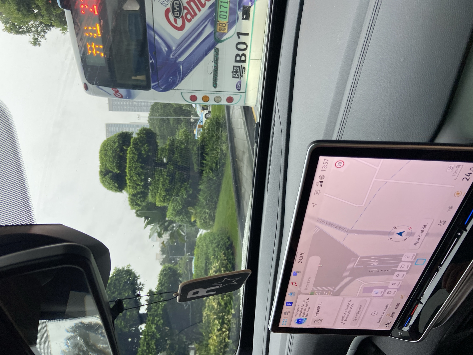 The image, taken from inside a vehicle, shows a traffic scene in Luohu District, China. The driver's side dashboard is visible, featuring a large touchscreen display with a navigation map. The map shows roads, including "Aiguo Road Side," and a blue arrow indicating the vehicle's current position. The exterior view through the windshield includes a bus with Chinese characters and the destination display showing "开往 2," indicating a route. Other cars and lush green trees are also visible in the background, suggesting an urban environment with significant greenery. A novelty air freshener hangs from the rearview mirror. The weather appears overcast.