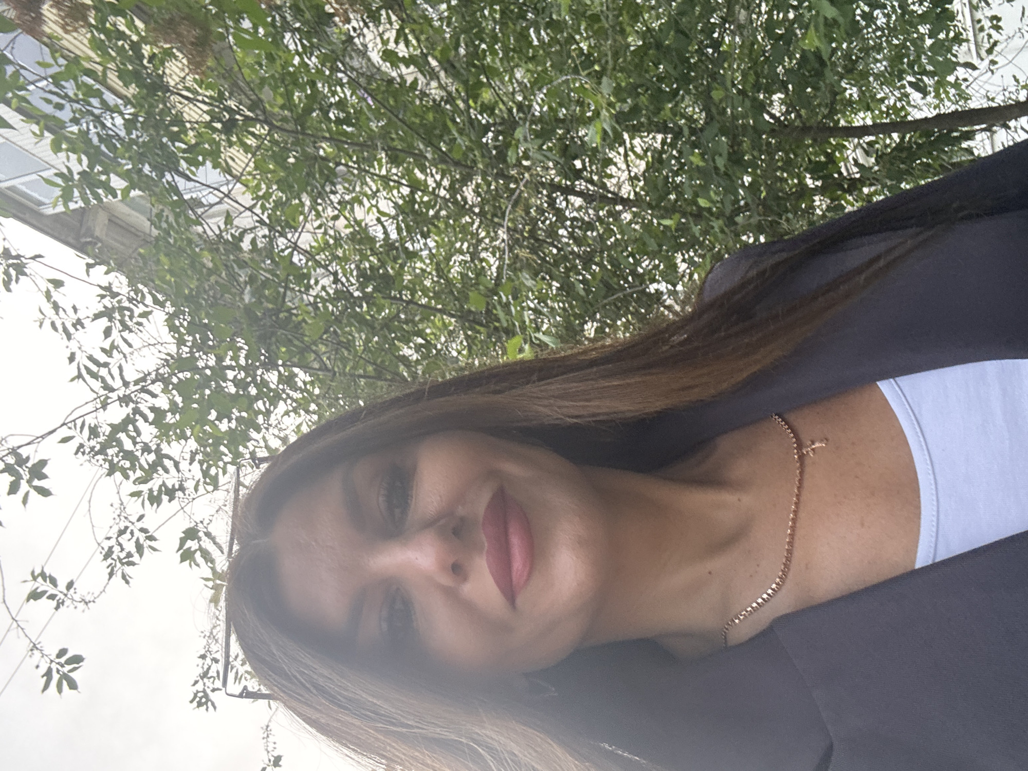A woman is posing outdoors, looking down at the camera. She has light brown hair and is wearing a black blazer over a light-colored top. A thin gold chain necklace is visible. Behind her are trees with green leaves, and in the upper left corner of the frame, part of a building with windows is visible. The location is potentially in a public or urban outdoor area, but the exact location in Chaikovsky, Russia, cannot be determined without further information or landmarks.
