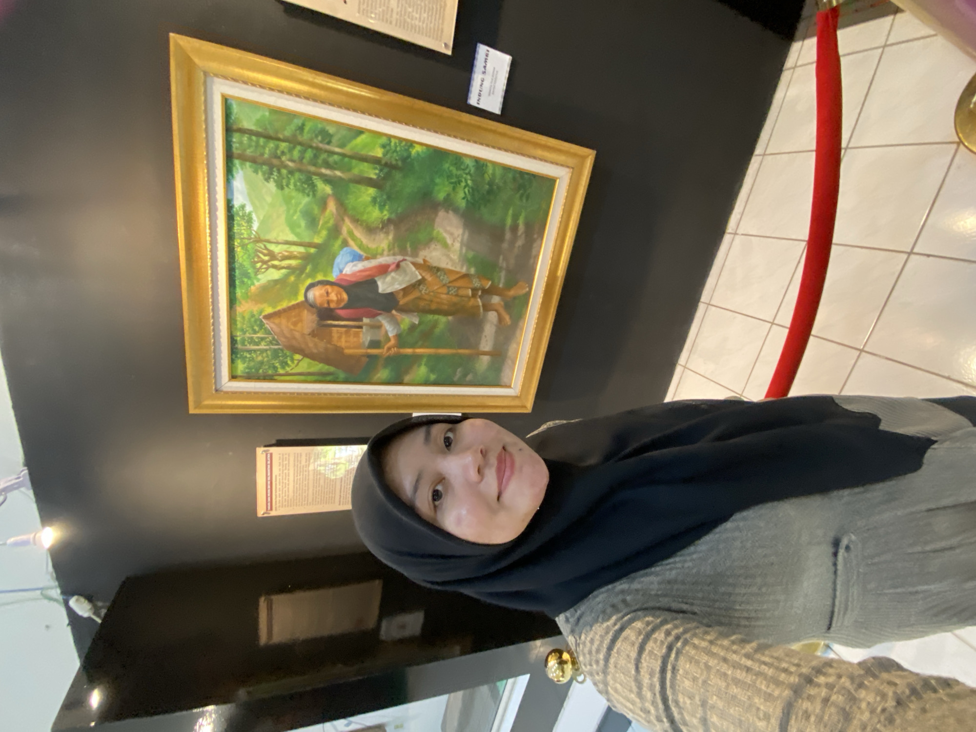 The image shows a woman wearing a black hijab and a light grey cardigan, taking a selfie in front of a painting. The painting, framed in gold, depicts an elderly woman walking on a dirt path through a lush green landscape with trees and a small hut. There is a red velvet rope barrier in the foreground. The background consists of dark walls and tiled flooring. A small label with text, including the word "INDUNG SAMS", is visible next to the painting, suggesting it might be displayed in a museum or gallery, possibly the Lambung Mangkurat Museum in Banjarbaru, South Kalimantan, Indonesia.