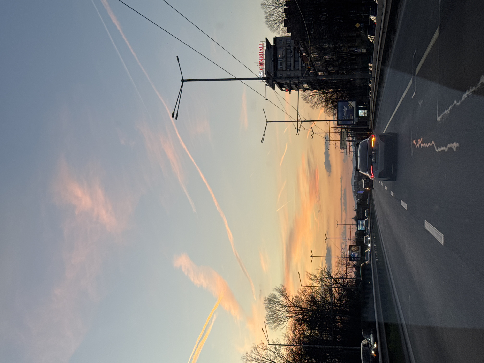 The image depicts a street scene in Sofia, Bulgaria at dusk. The sky is a gradient of light blue to orange-pink, marked with prominent contrails. A line of streetlights extends along the road, their poles and wires visible against the sky. On the right side of the image, a multi-lane road is visible with cars traveling along it. Buildings line the roadside, including one with "GENERALI" in red neon lettering on top. Bare trees stand between the road and the buildings. The foreground is dominated by the road surface, with painted lane markings.