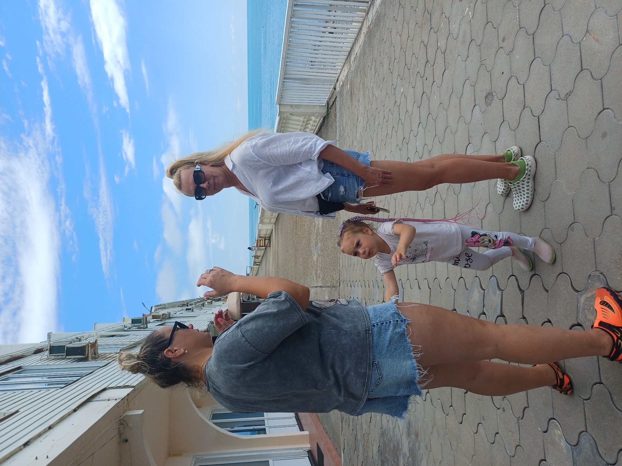 Two women and a young girl stand on a paved walkway beside the sea. The woman on the left wears a gray t-shirt, denim shorts, and sunglasses. Her hair is pulled back in a bun. The woman on the right wears a white open-collared shirt, denim shorts, sunglasses, and a fanny pack. Her blonde hair is in a ponytail. The girl, positioned between the two women, wears a white t-shirt and leggings. She has purple braids. The walkway is paved with interlocking gray tiles. A white railing separates the walkway from the sea, which is visible in the background under a blue sky with white clouds. The scene appears to be a promenade in Sochi, Russia.