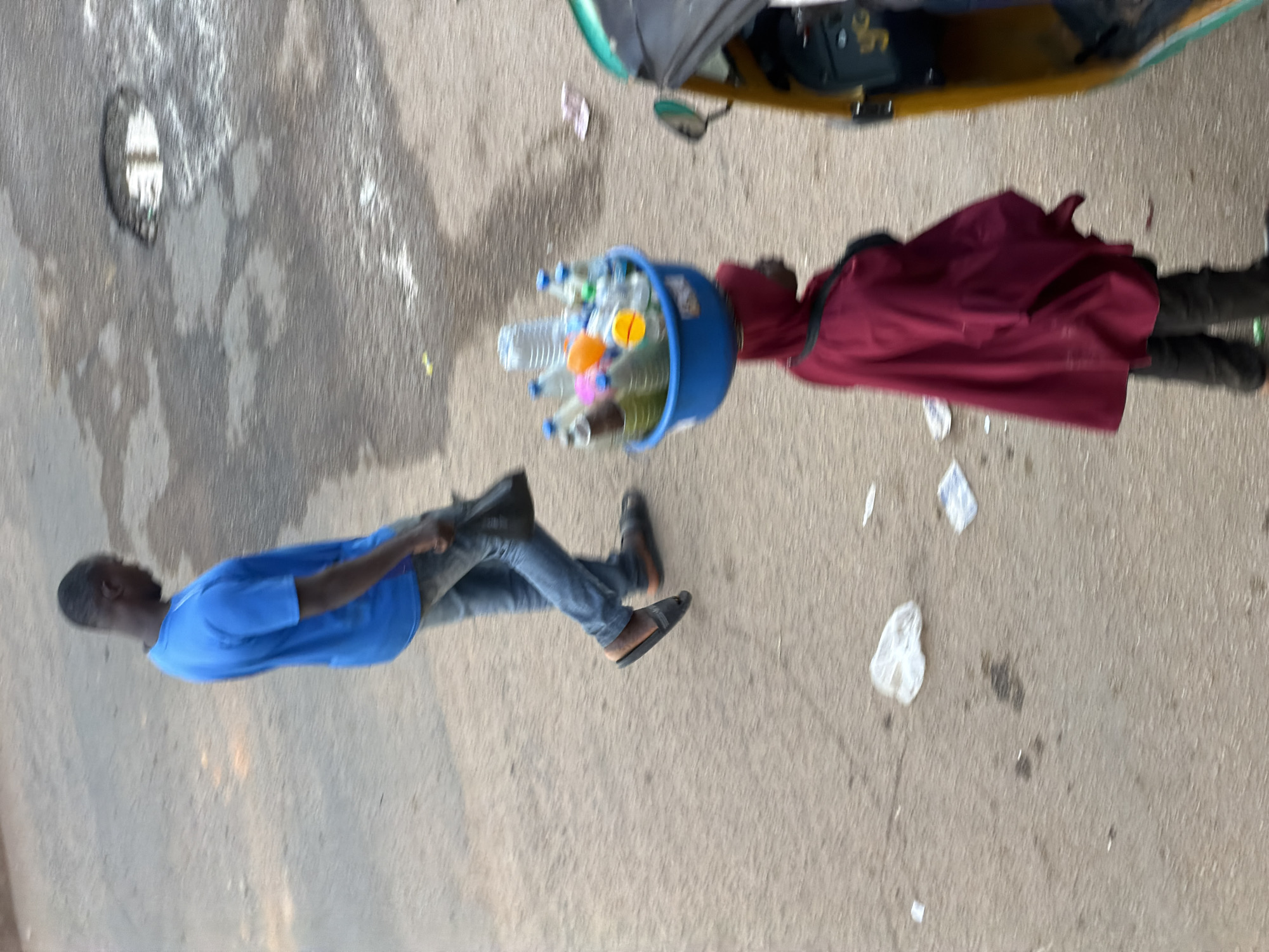 In Bukuru, Nigeria, on a paved surface, a man in a blue t-shirt and jeans walks to the left, carrying a black bag in his right hand. To his right, a person wearing a burgundy hooded garment and dark pants walks to the right, balancing a blue bucket filled with various bottles of liquid on their head. A yellow and green auto-rickshaw is parked to the right of the person with the bucket. The ground is rough and uneven, with scattered debris and a puddle of water.