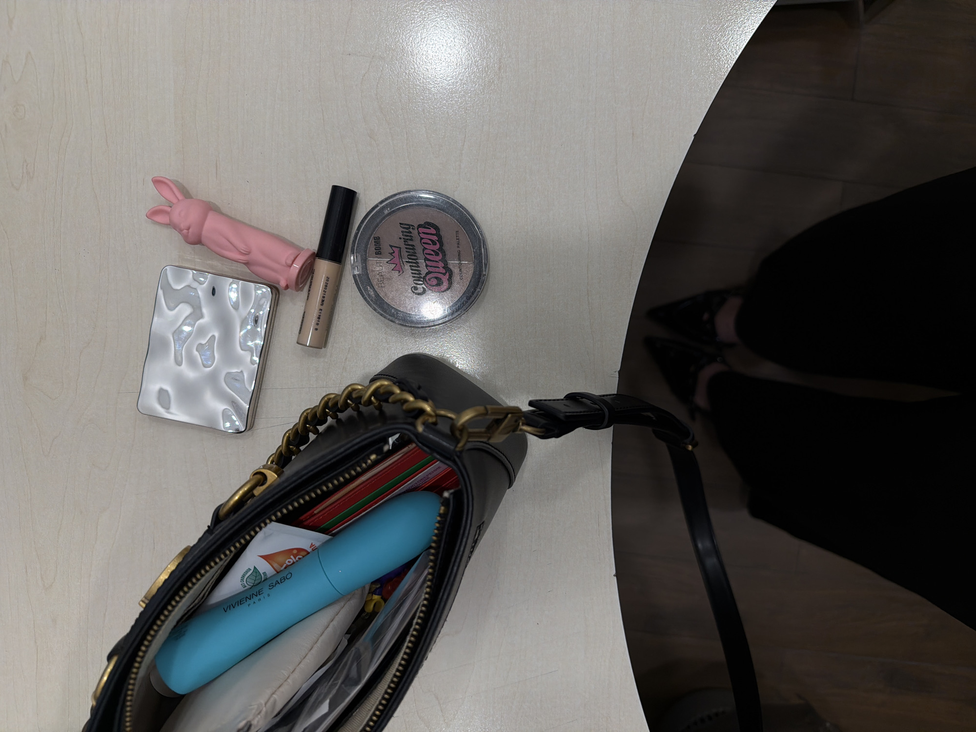 The image shows a light-colored surface with a black purse and various items scattered around it. These items include a pink bunny-shaped object, a silver compact, a concealer, and a contouring palette. The purse contains a blue mascara tube and other items. A portion of a person's legs and shoes are visible in the lower right corner. The location, Izhevsk, Russia, cannot be determined from the given image.
