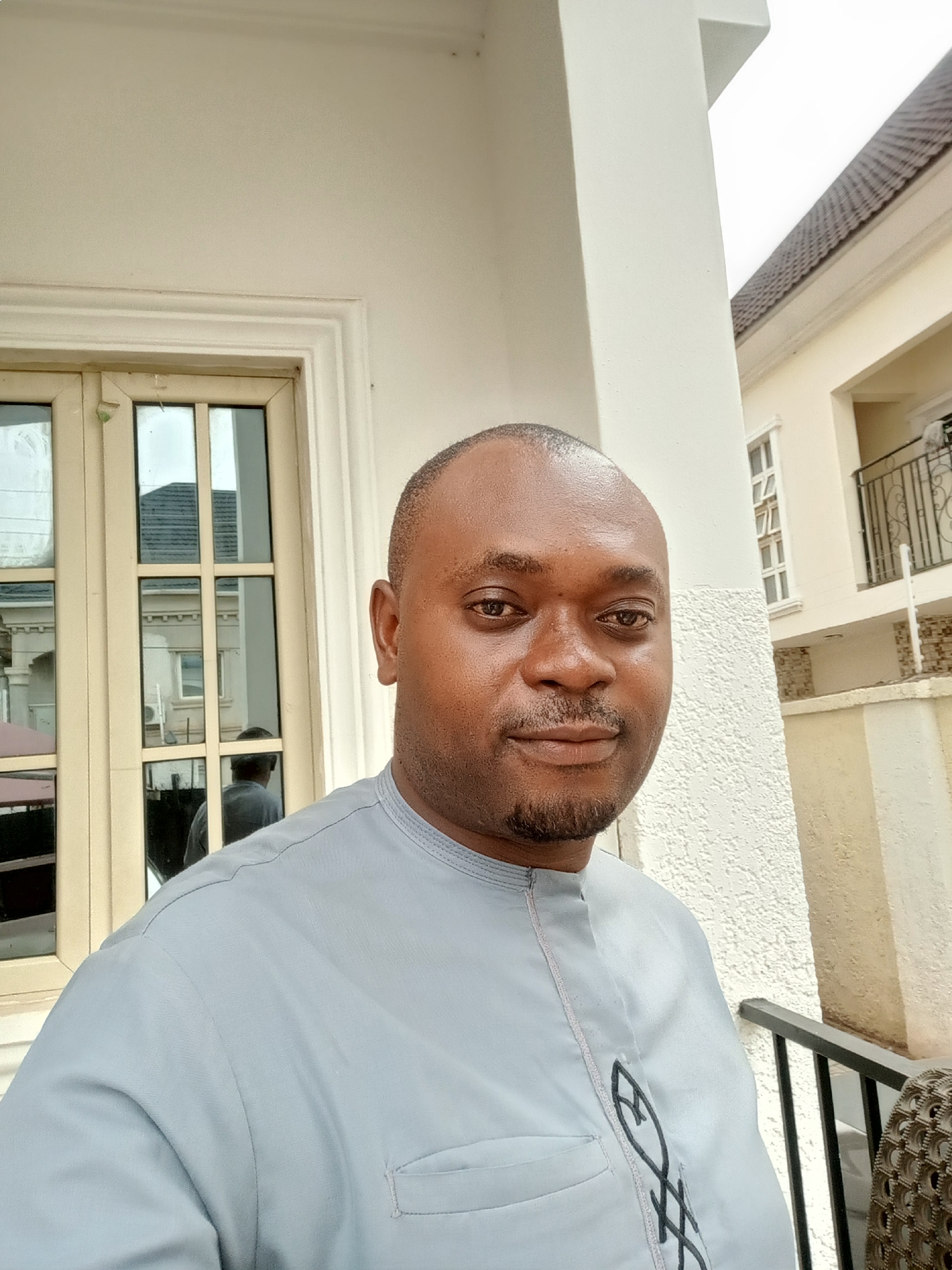 A medium shot depicts a man standing outdoors in what appears to be a residential area in Gwarinpa, Nigeria. The man has dark skin, a shaved head with some stubble, and a neatly trimmed beard. He is wearing a light gray traditional garment with an embroidered design on the front. Behind him are architectural elements of buildings. A window with multiple panes reflects part of another building. A column and sections of a balcony or railing are visible, suggesting the presence of multiple structures. The overall setting implies a relatively affluent neighborhood.