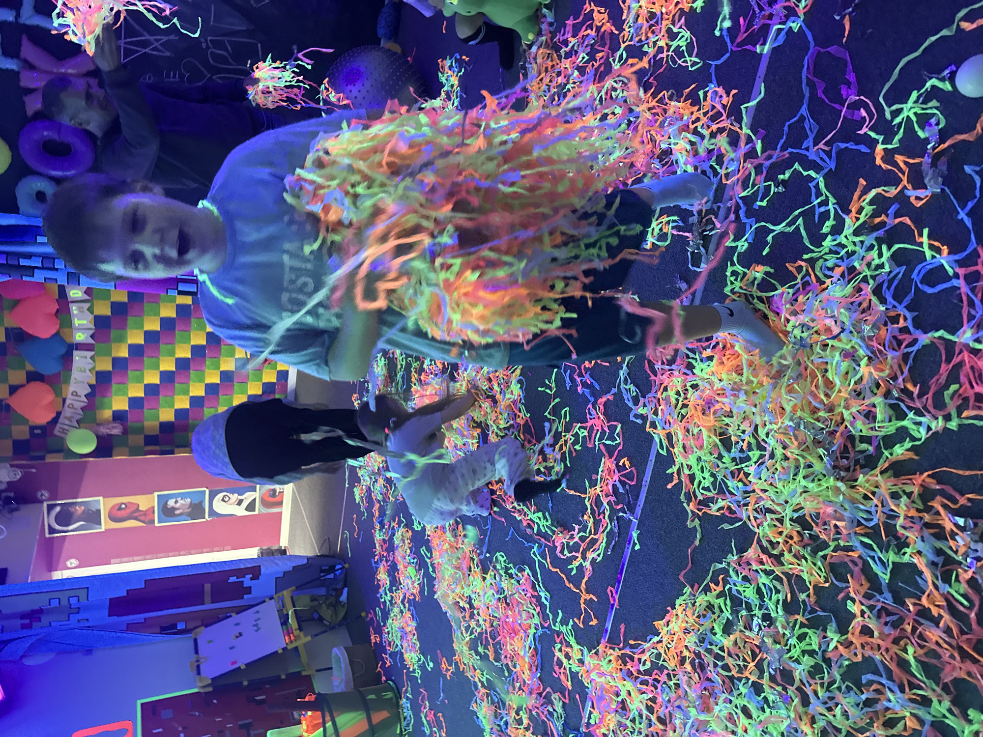 The image depicts an indoor setting, possibly a party or event space in an unknown city. Fluorescent confetti lit by blacklights covers the floor. A young boy in a light blue t-shirt is holding a large, colorful confetti pom-pom overhead, with a wide-mouthed expression, perhaps yelling or laughing. He is standing amidst the confetti. A child is kneeling to the right, also interacting with the confetti on the floor. Additionally, there is another child in the background to the left, behind the boy, holding what appears to be another confetti pom-pom.
The backdrop includes a checkered wall design with balloons and a "Happy Birthday" banner. A series of framed pictures, possibly superhero characters, adorn a pink wall in the background. Play equipment and other party-related items are scattered around the room.
