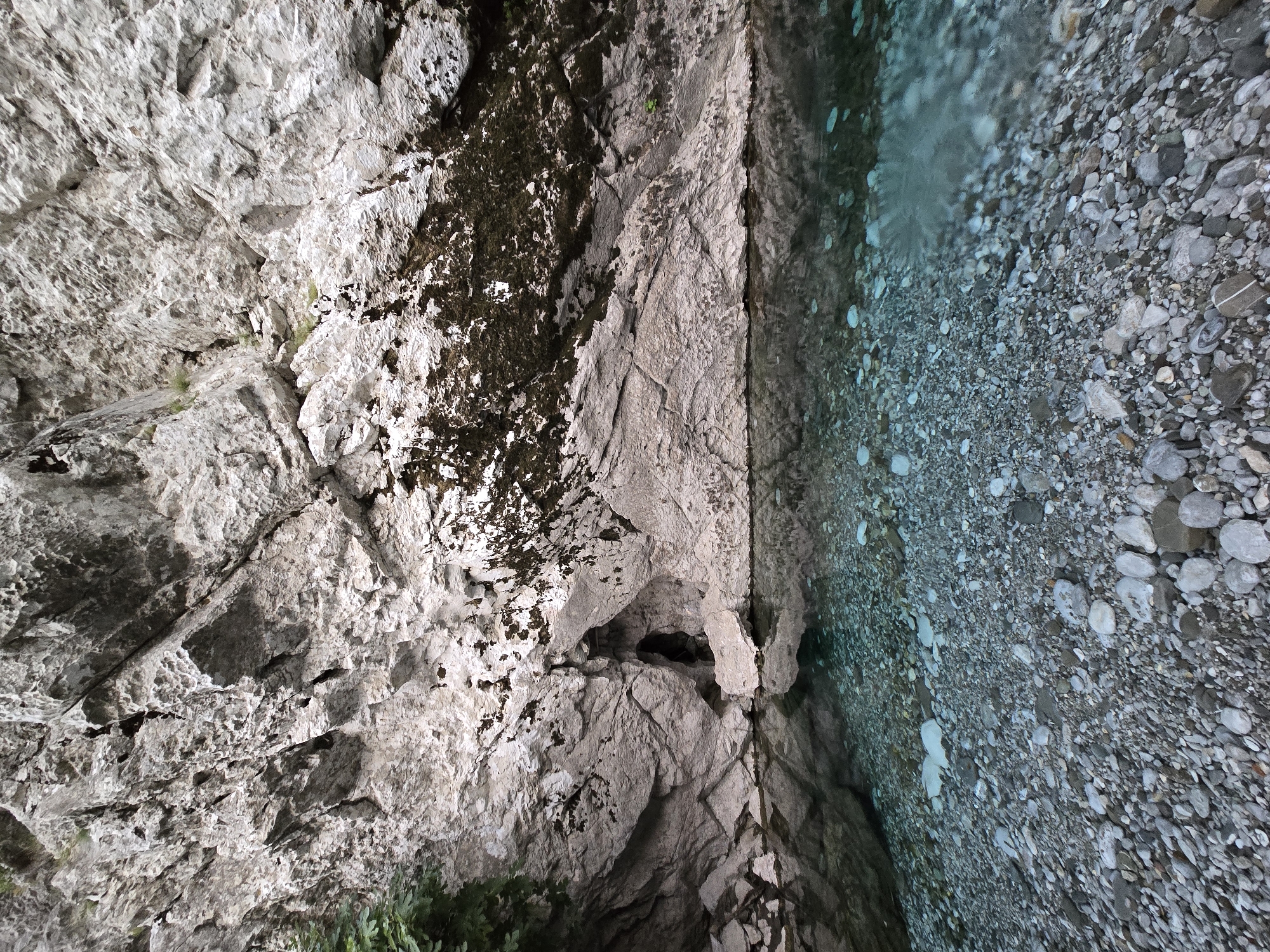 The image shows a clear, shallow body of water with a rocky bed. The water reflects a large, rugged, light-colored rock face that dominates the background. The rock face has various textures, including rough, weathered surfaces and darker patches, possibly moss or algae. A small, dark opening, resembling a cave or crevice, is visible within the rock face, just above the waterline. The water itself is transparent, revealing the numerous smooth, rounded pebbles and stones that make up the streambed. The scene appears to be a natural landscape, likely in a mountainous or canyon area, possibly near Bioče, Montenegro, given the characteristic rocky terrain and pristine water often found in such regions. There are no people or animals visible in the image.