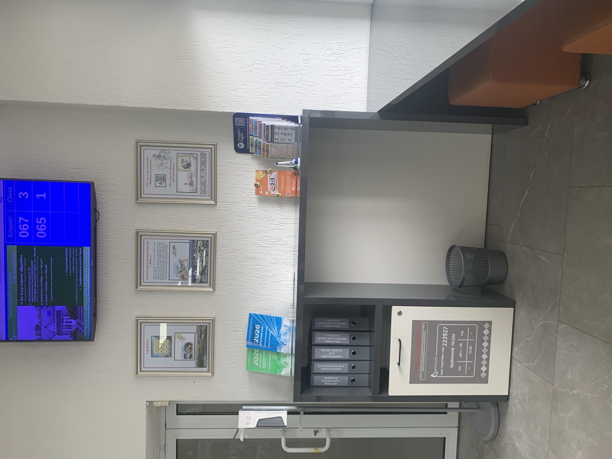 The image depicts an indoor scene, likely a service or information center, with a digital display screen prominently featuring the text "ПРИГЛАШАЕМ В КОМАНДУ" and "РУП «БЕЛПОЧТА»". The screen displays customer queue numbers, with "067" and "065" listed under "Клиент" (Client) and "3" and "1" under "Окно" (Window). Below the screen, on a wall, are three framed exhibits. The left exhibit features a picture of a house and possibly postage stamps. The middle exhibit displays text and images related to Grodno and potentially Belarusian history. The right exhibit contains text and images, with one image appearing to be a portrait and another a city panorama, possibly relating to Belarusian heritage or culture.
In the foreground, a grey desk with a dark countertop holds several items. On the left side of the desk, there are two blue binders labeled "2026". To the right of these binders is a set of six grey binders in a shelf unit. Below the binders, a white and grey cabinet is visible, displaying a sticker with information about "НАШ ПОЧТОВЫЙ КОД 223927" and "ВЫХАННЕ ПІСЕМ" (outgoing mail). A silver-colored trash can is situated to the right of the cabinet. Further to the right on the desk are various brochures and leaflets, including those from "СПОРТПАРИ", one with "3+3" visible.
A dispenser for hand sanitizer is attached to a doorframe on the far left of the image. The floor is tiled with a marble-like pattern. The walls are textured and painted a light color. Two orange upholstered seats are visible on the right side of the image, partially under the desk. The scene appears to be well-lit, suggesting an interior space.
The presence of "РУП «БЕЛПОЧТА»" on the display screen indicates this is a branch of the Belarusian Post. The inclusion of Belarusian cultural and historical exhibits suggests a local context within Belarus. The text "223927" and "Капыль, Беларусь" (Kapyĺ, Belarus) on one of the brochures or signs, though not fully legible, strongly suggests the location is Kapyĺ, Belarus.
