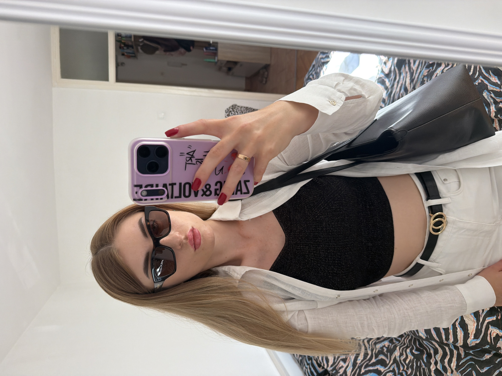 This is a portrait-oriented, vertical photograph taken of a woman posing for a mirror selfie. The woman has long, blonde hair, is wearing sunglasses with black frames, a black knit top, a white unbuttoned shirt over the top, and white pants with a black belt featuring a gold double-ring buckle. She is holding up a purple phone with a camera lens visible. Her nails are painted red, and she is wearing a gold ring on her left ring finger. A black handbag is slung over her shoulder. The background appears to be an indoor setting, with a white wall and a mirror reflecting part of a room with furniture. The overall lighting suggests it might be daytime. The woman's expression is neutral, and the scene is casual. Text is visible on the phone case, which appears to be in black font.