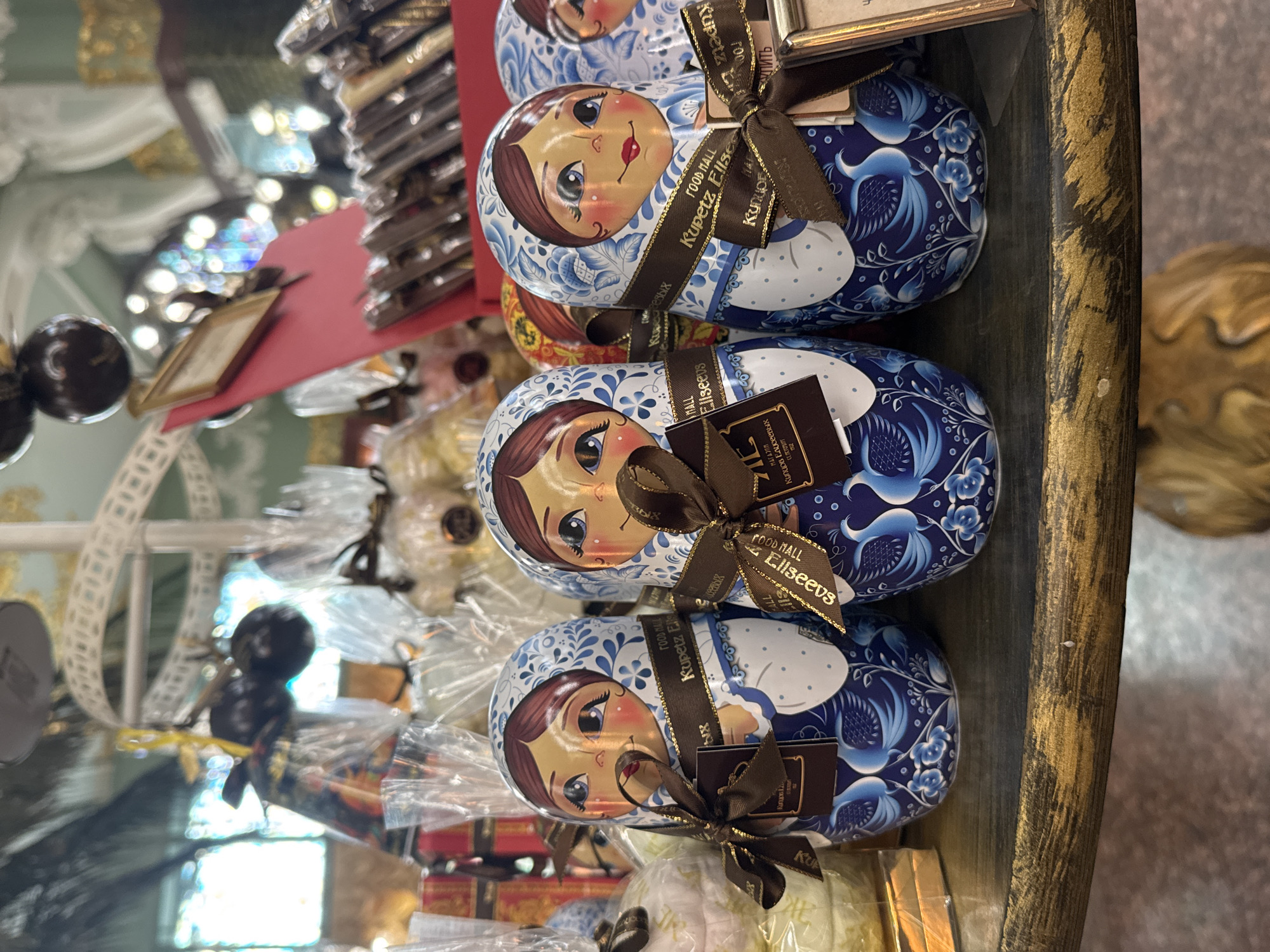 The image shows a display of Matryoshka doll-shaped tins, each adorned with a traditional blue and white floral design and featuring the face of a Russian woman. Each tin is wrapped with a brown ribbon and a tag that reads "Kupetz Eliseevs," indicating the location of the display as the Eliseev Emporium in Saint Petersburg, Russia. The scene is within a shop, with other packaged goods visible around the matryoshkas.