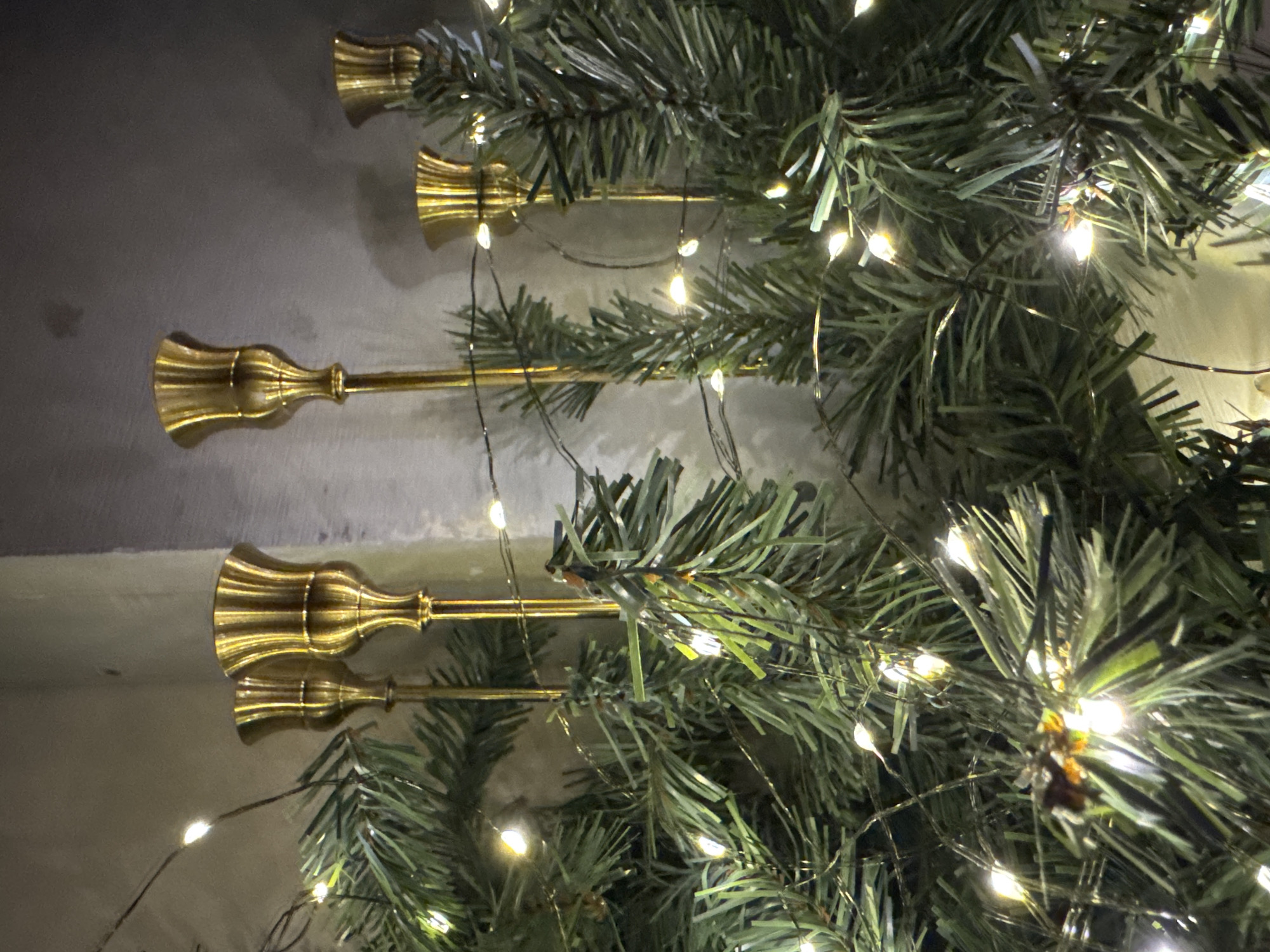 The image shows a close-up of a decorative arrangement featuring several brass-colored candle holders interspersed with artificial pine branches and illuminated by a string of white fairy lights. The candle holders have fluted tops and slender stems. The pine branches appear realistic with green needles. The fairy lights are small and bright, casting a warm glow. The background is a textured, neutral-colored wall, likely concrete or plaster. The overall impression is one of festive, indoor decoration. The location in Steklyannyi, Russia, is not visually represented in the image itself, as the scene is entirely focused on the decorative objects.