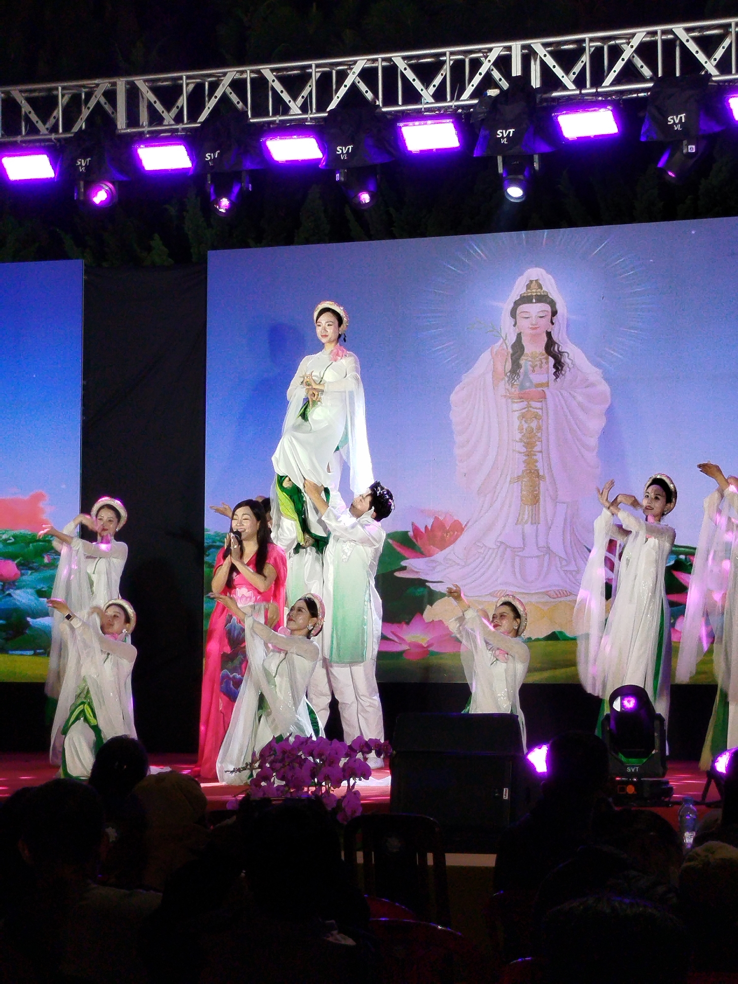 Onstage, a woman in a red dress with floral accents sings. Surrounding her are dancers in white traditional dresses, performing in front of a screen displaying an image of a deity. Above the stage, lighting fixtures illuminate the performance. The scene is likely set in Da Lat, Vietnam, given the cultural references in the performance. A crowd of people are seated in the foreground watching the show.