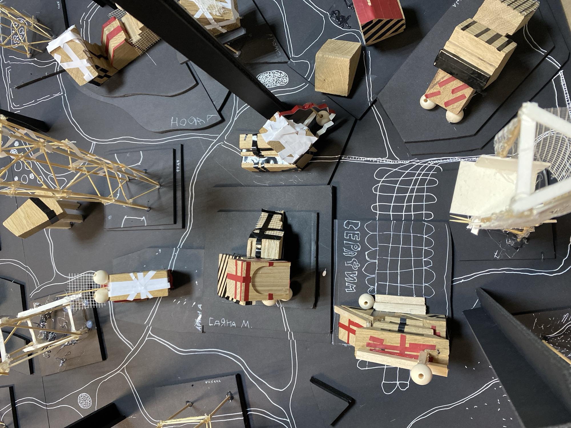 The image shows an architectural model of a possible urban plan for an area in Moscow, Russia. The model uses a black base on which a road network is sketched with white lines. Several small building models are scattered across the base. These buildings are constructed from various materials like wood blocks, sticks, and paper. Some buildings are decorated with red or black stripes and white tape. Handwritten labels in Cyrillic are placed near some of the building models, likely indicating location or building type. The scene suggests a design study or presentation of architectural proposals for urban development.