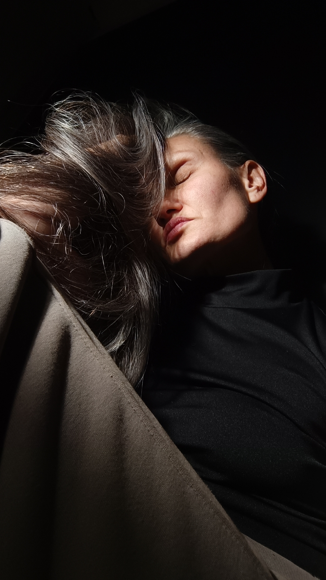 A woman with graying hair lies with her eyes closed, her face partially illuminated by a beam of light. She is wearing a black top. A light brown fabric covers part of the left side of the image. The background is mostly dark. The location is unknown but speculated to be in the city of Lermontov, Russia.