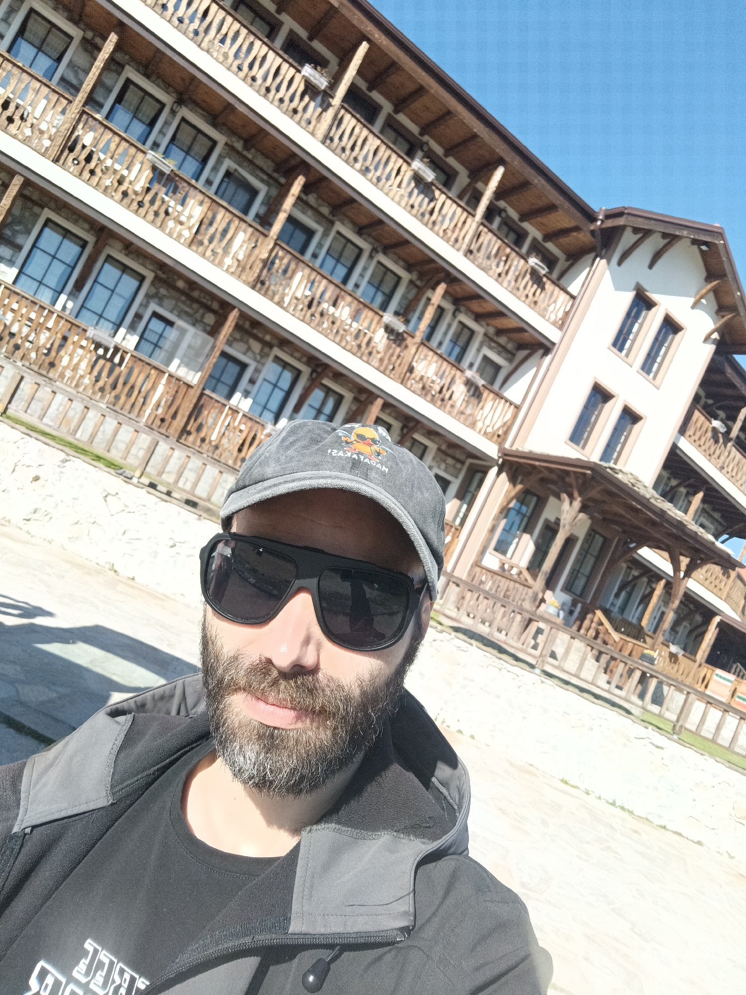 A man with a beard and sunglasses is taking a selfie in front of a building. The building has multiple floors, balconies, and windows. The building's architecture is a combination of stone and wood. The sky is clear and blue. The location is likely Vrata, Bulgaria, based on the man's clothing logo of the Vratsata.