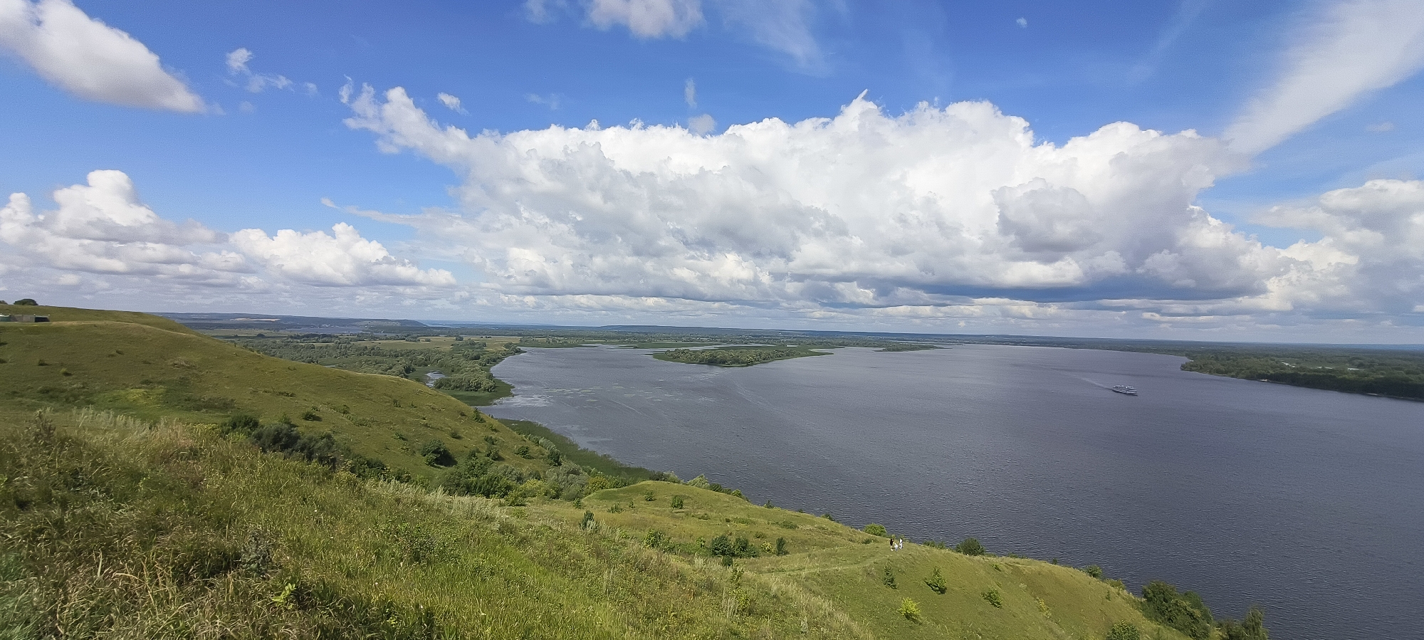This is a panoramic, eye-level, outdoor, daytime photograph taken from a grassy hillside overlooking a wide, calm river under a partly cloudy blue sky. The foreground is dominated by the lush green grass of the hillside, with some patches of taller weeds and scattered small trees and bushes. The hillside slopes down towards the river, with a winding path visible on its surface. In the mid-ground, the river curves and widens, with lush green, tree-covered banks on either side. A small white ship is visible on the river, moving away from the viewer and towards the right side of the frame. In the background, the river continues to widen, with a flat, green landscape stretching to the horizon, dotted with trees and appearing to have some buildings in the distance. The sky is a vibrant blue, filled with large, fluffy white clouds. The overall scene depicts a serene and expansive natural landscape.