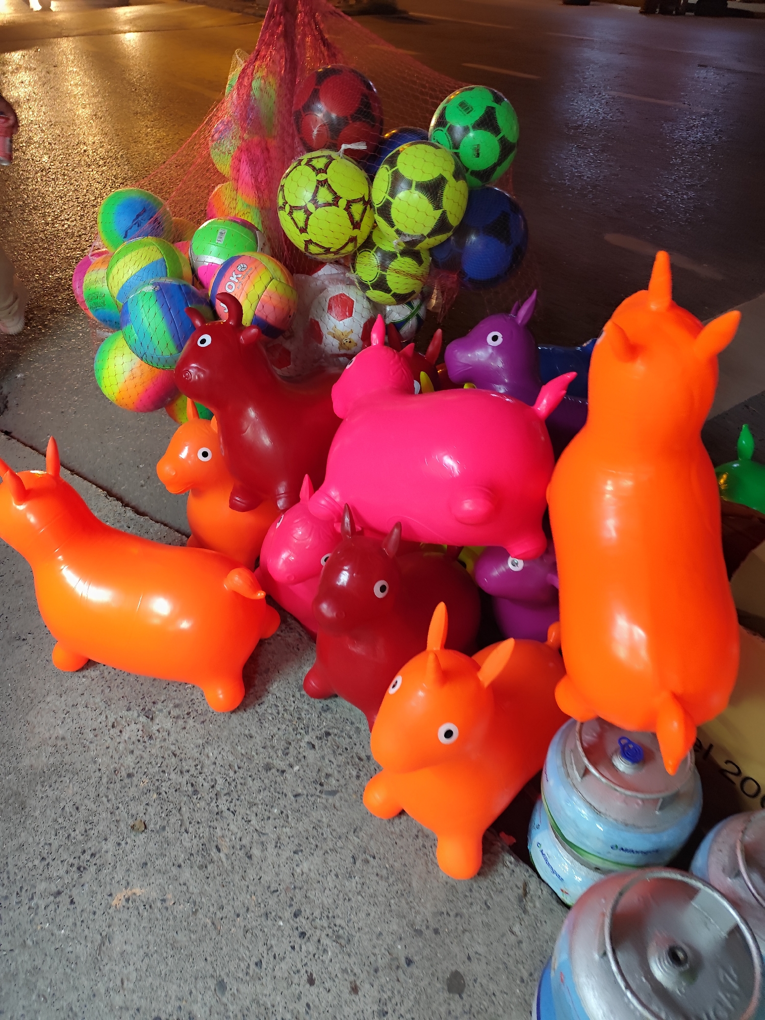 Captured at night on what appears to be a wet street in Sarıyer, Turkey, the image showcases a collection of colorful toys arranged on a concrete surface. Several inflatable, ride-on animal toys, predominantly in shades of orange, red, pink, and purple, are prominently displayed. Alongside the animals, a net holds a variety of colorful balls, including soccer balls and other designs. Cylindrical objects, potentially containers or canisters, are positioned near the toys, partially obscured. The scene suggests a street vendor or market stall selling toys. The background is dark, illuminated by streetlights and reflecting off the wet pavement, which further suggests the location is outdoors and in an urban setting. A person's leg is partially visible at the edge of the image, indicating the presence of people in the vicinity.