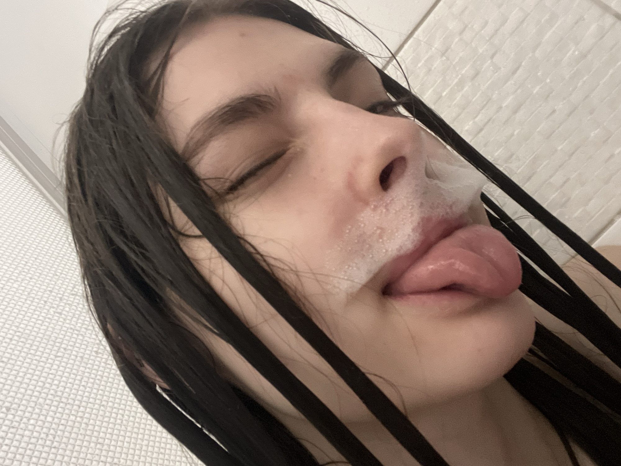 A close-up, angled shot features a young woman with dark, wet hair plastered to her face. Her eyes are closed, and her tongue is extended, covered in a bubbly, white substance. Some of the same substance is smeared around her upper lip and nose. The background shows white, textured wall tiles. No information suggests this image was taken in Saint Petersburg, Russia.