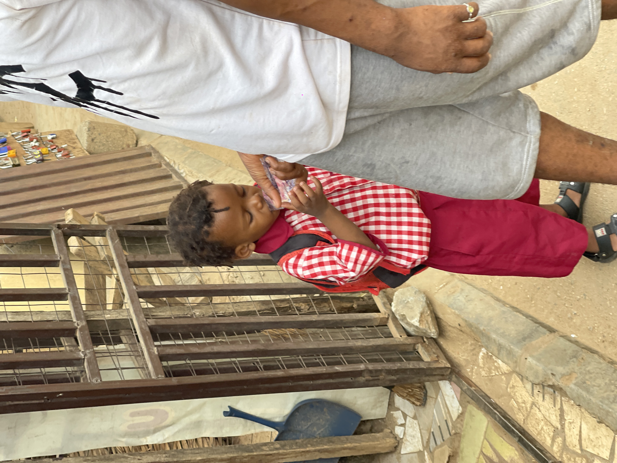 A child wearing a red and white checkered shirt and red bottoms is drinking from a small, clear plastic bag held in their hands. An adult wearing a white shirt and grey shorts is standing nearby. The scene appears to be outdoors, in Kuchingoro, Nigeria, next to a metal grate and a dustpan. A stone border and a partially visible tiled mosaic are also in view.