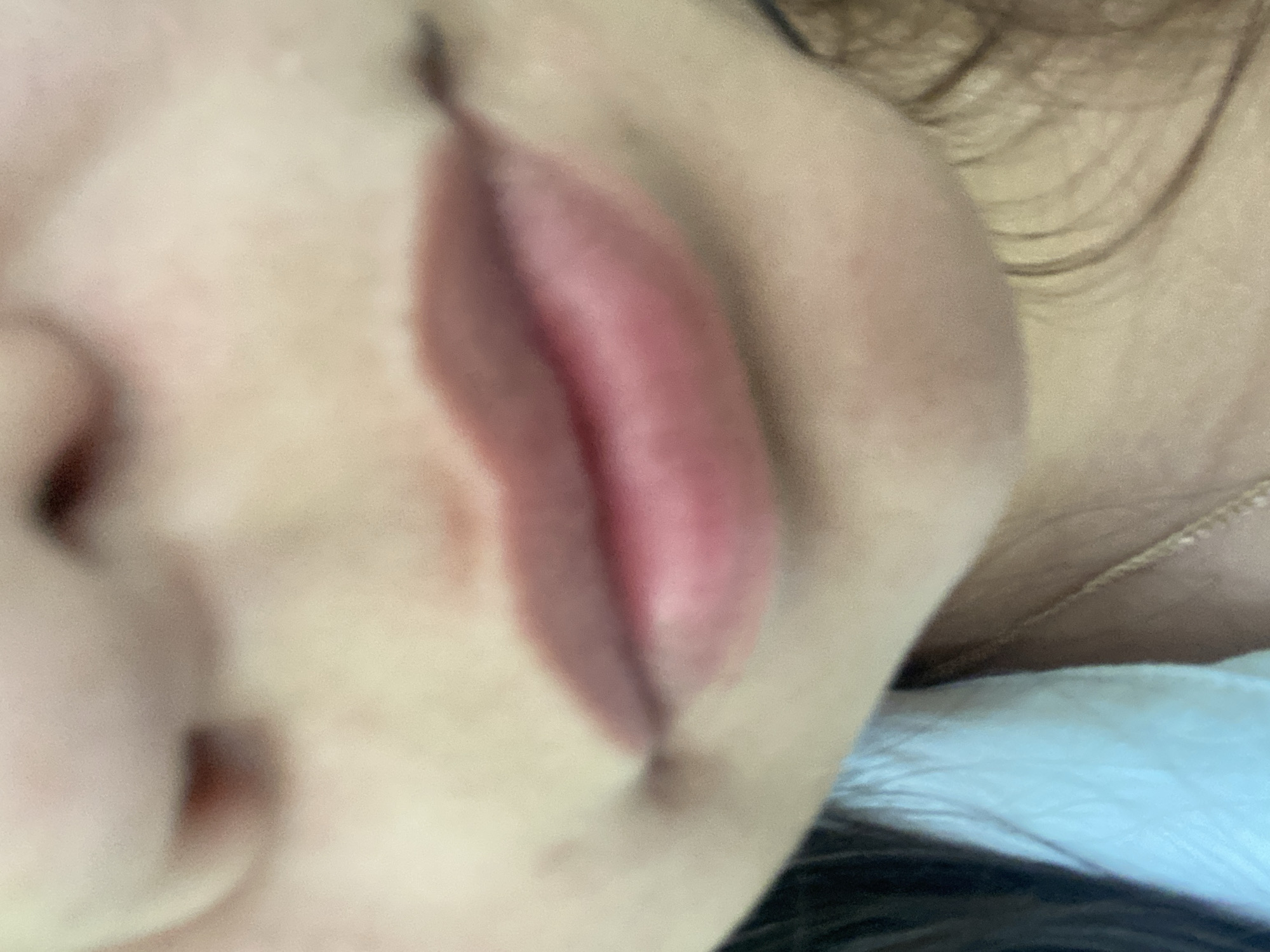 This is a close-up, slightly out-of-focus image of a person's mouth and lower face. The lips are slightly parted and appear to have a pink or reddish tint, possibly from lipstick or natural coloring. There are some freckles or blemishes visible on the skin. The person is wearing a light blue garment, possibly a shirt or blouse, and a thin gold necklace can be seen. The background is blurry and does not provide any clear environmental details. The image does not contain any information about the city of Tyulyugul, Russia.