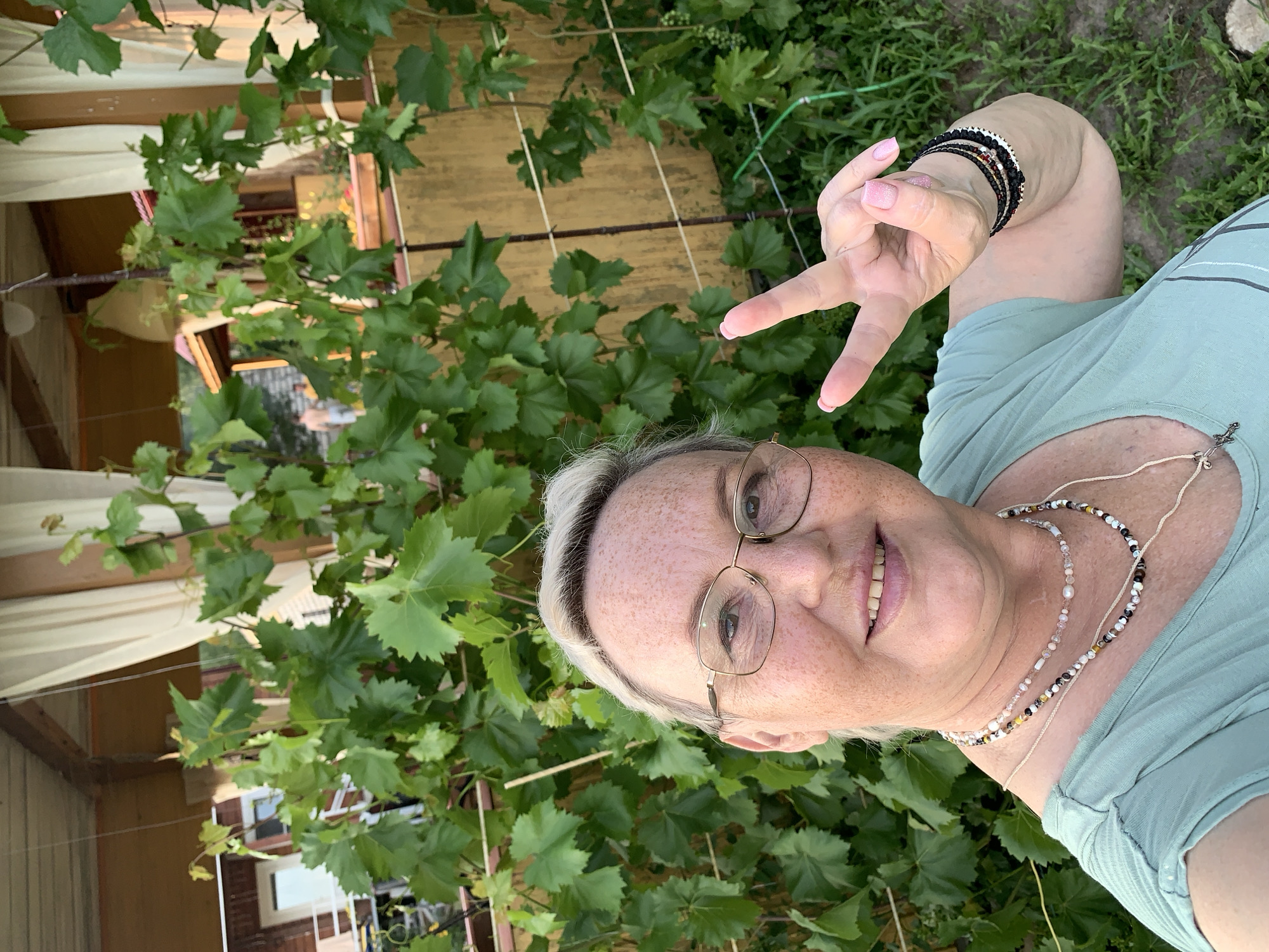 In a selfie taken outdoors in Surovatikha, Russia, a woman with fair skin, freckles, and blonde hair pulled back is wearing glasses and a light green t-shirt. She has on multiple beaded necklaces and bracelets on her right wrist. Her right hand is raised, forming a peace sign. Behind her, lush green grape leaves from a vine create a natural backdrop, partially obscuring a weathered yellow wall and a wooden structure with draped curtains. In the distance, a small building with a red roof and two people are visible. The ground is covered in grass and other greenery.