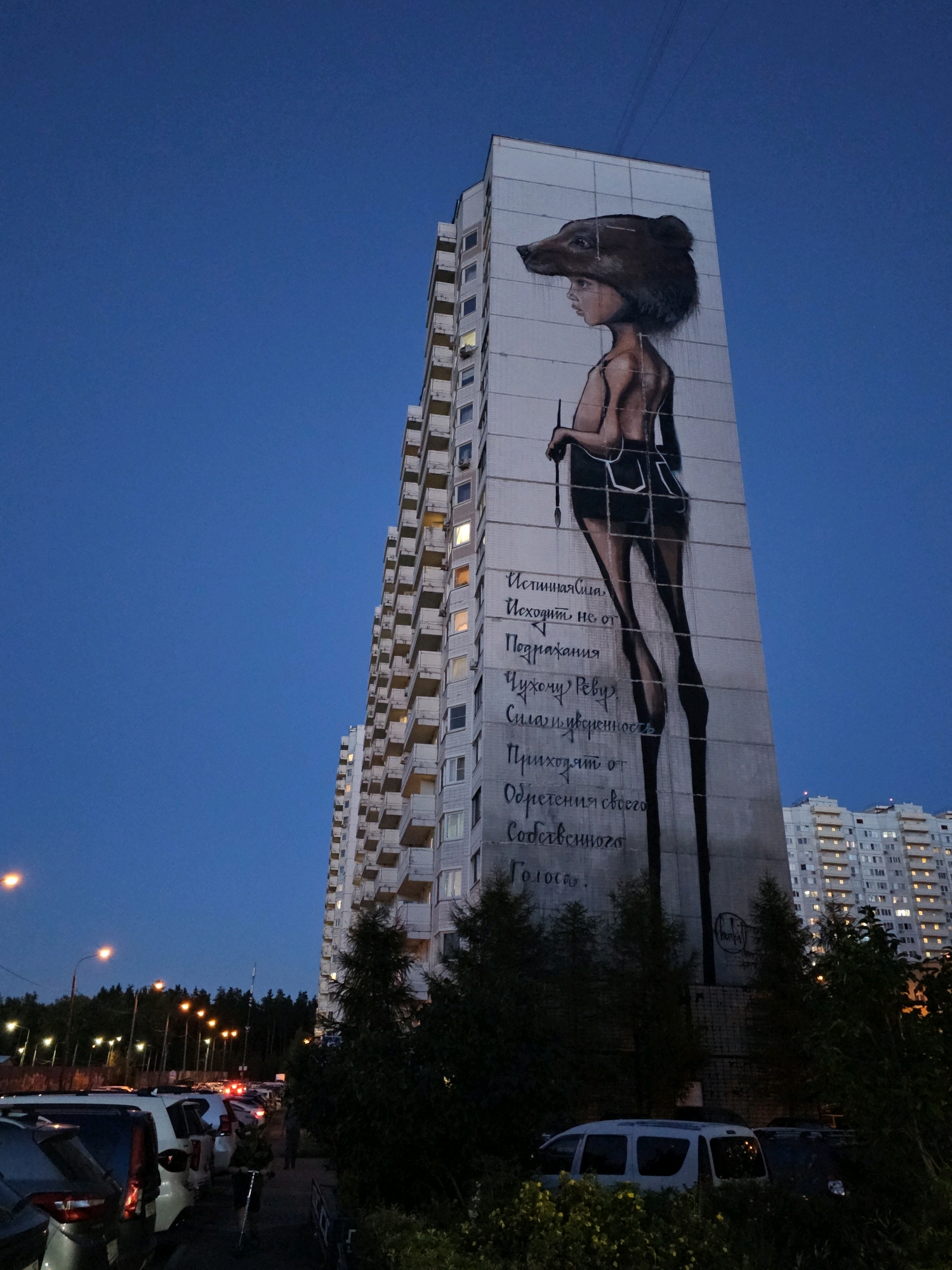 A tall, multi-story apartment building features a large mural of a person with a bear head, viewed in profile, facing left. The person is depicted with a thin body, dark shorts, and long legs, holding a stick. Russian text is painted vertically on the building's facade next to the mural. The scene is set at dusk, with the sky a deep blue. Streetlights illuminate a row of parked cars on the left side of the image, and more apartment buildings are visible in the background to the right. Trees and foliage are present in the foreground and around the base of the building. The location is identified as Odintsovo, Russia.