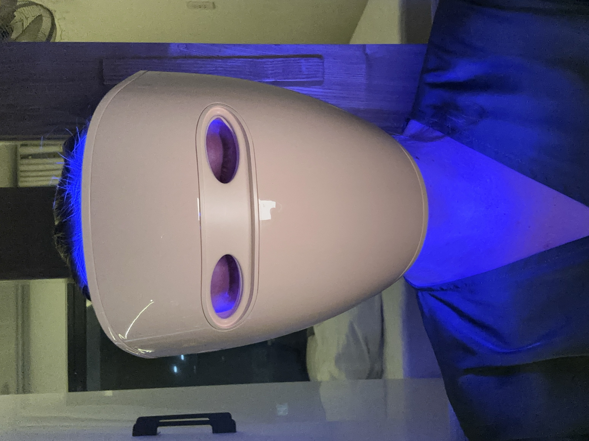 An individual is wearing a pink LED facial mask that emits blue light. The mask covers the entire face and has eye openings through which the person's eyes are visible, illuminated by the blue light. The individual is also wearing a dark blue robe or garment that is illuminated by the blue light from the mask. The background appears to be an indoor setting with a bed and what seems to be a window or glass partition. The overall lighting is dominated by the blue light from the mask. There is no identifiable information in the image to suggest a specific location in Đà Nẵng, Vietnam.
