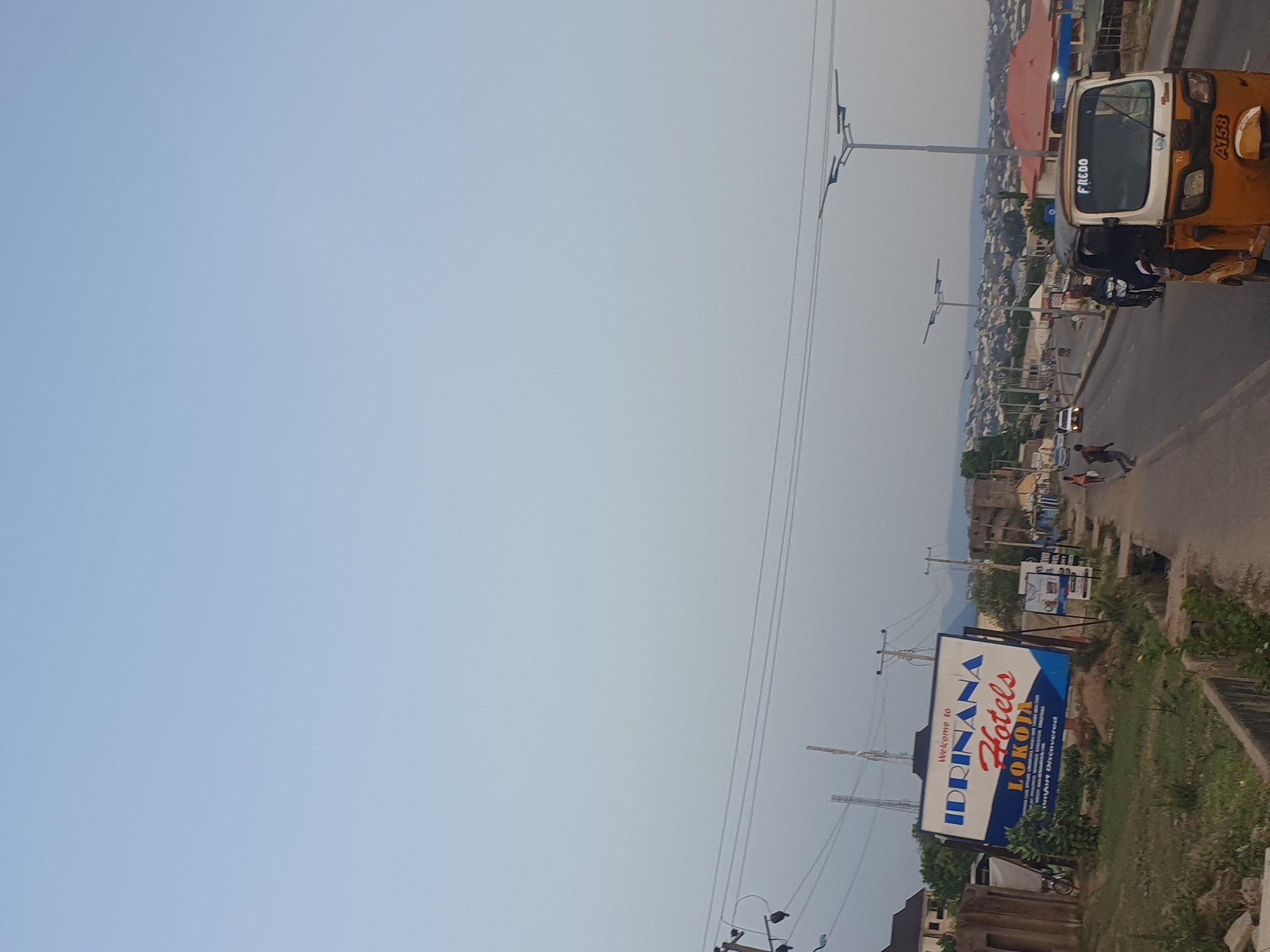 The image depicts a roadside scene in Lokoja, Nigeria. The sky is a pale blue, suggesting either early morning or late afternoon. An "Idrinana Hotels Lokoja" sign is prominent on the right side of the image, indicating the location. A three-wheeled vehicle (auto rickshaw), locally known as a "keke napep" is seen on the road, with the name "Fredo" printed on the windshield, and the registration number "AT53" on the side. Two people are walking along the side of the road. Electrical poles and wires are visible overhead. The terrain includes green vegetation on the road edges, indicative of a tropical environment. In the background, a cityscape unfolds with buildings of varying heights and styles, revealing the urban fabric of Lokoja.