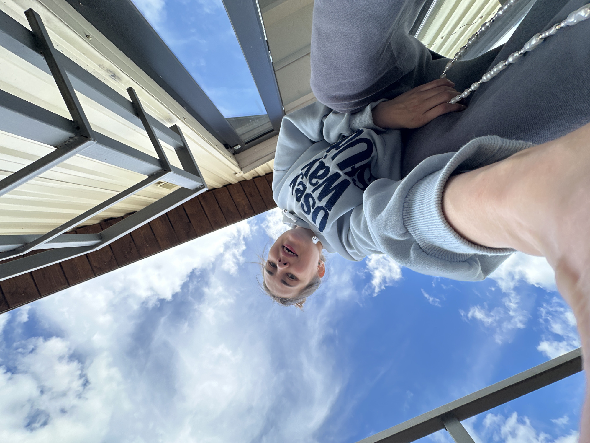 A young person is pictured from a low angle, with the sky visible behind them. The person is wearing a light-colored hooded sweatshirt with dark lettering. A string of pearls or beads is visible around their neck. The environment includes architectural elements such as beams and roof structures. The image was taken in Ilyinsky, Russia.