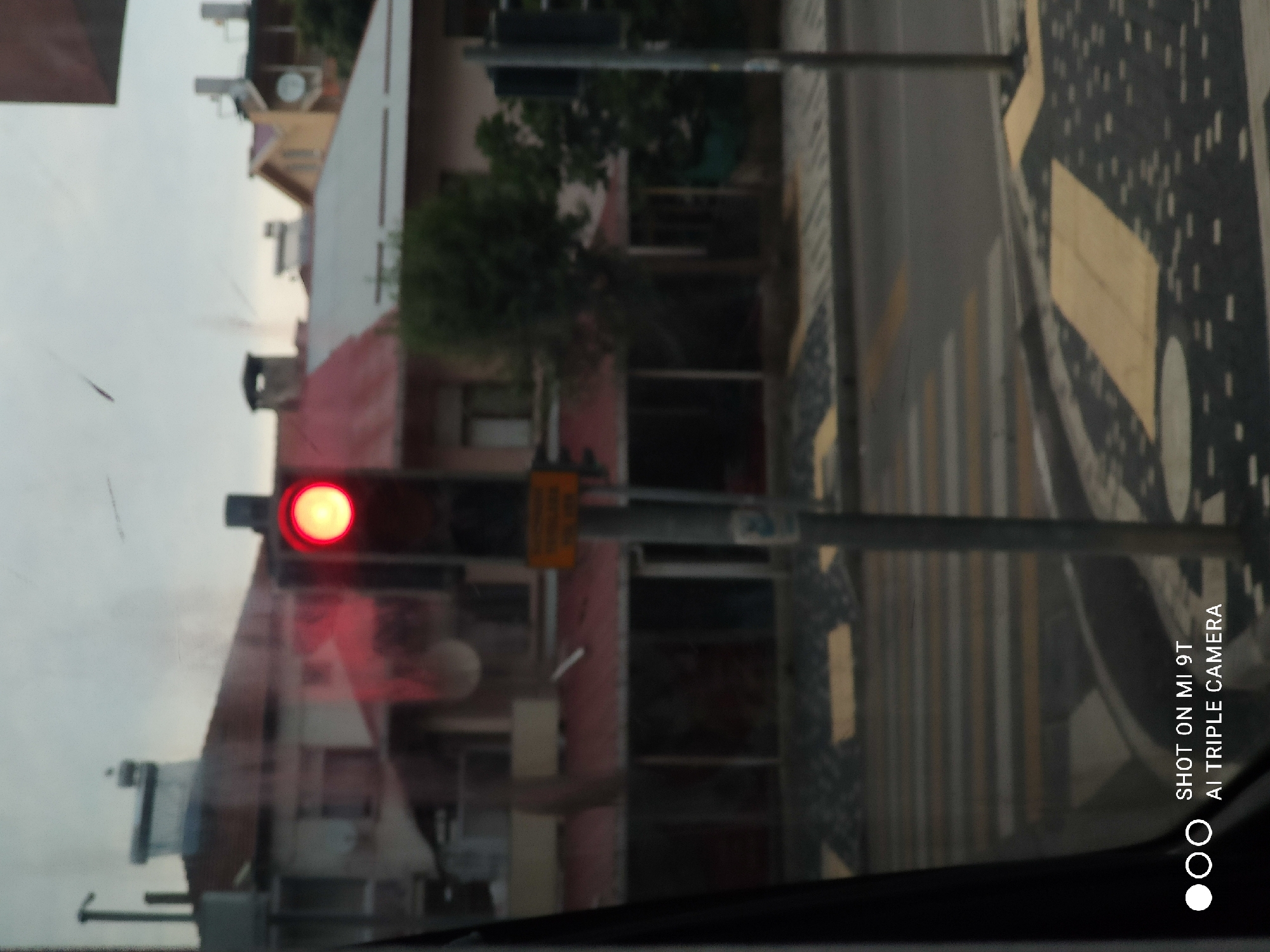 This image depicts a traffic light with a red signal illuminated, positioned in front of buildings and a street. The scene appears to be a city environment, possibly in Trabzon, Turkey, given the context. The buildings have red roofs and various architectural features, including chimneys and satellite dishes. A crosswalk is visible on the street, with yellow markings. The lighting suggests an overcast or cloudy day. The image also includes a watermark at the bottom left indicating "SHOT ON MI 9T AI TRIPLE CAMERA".