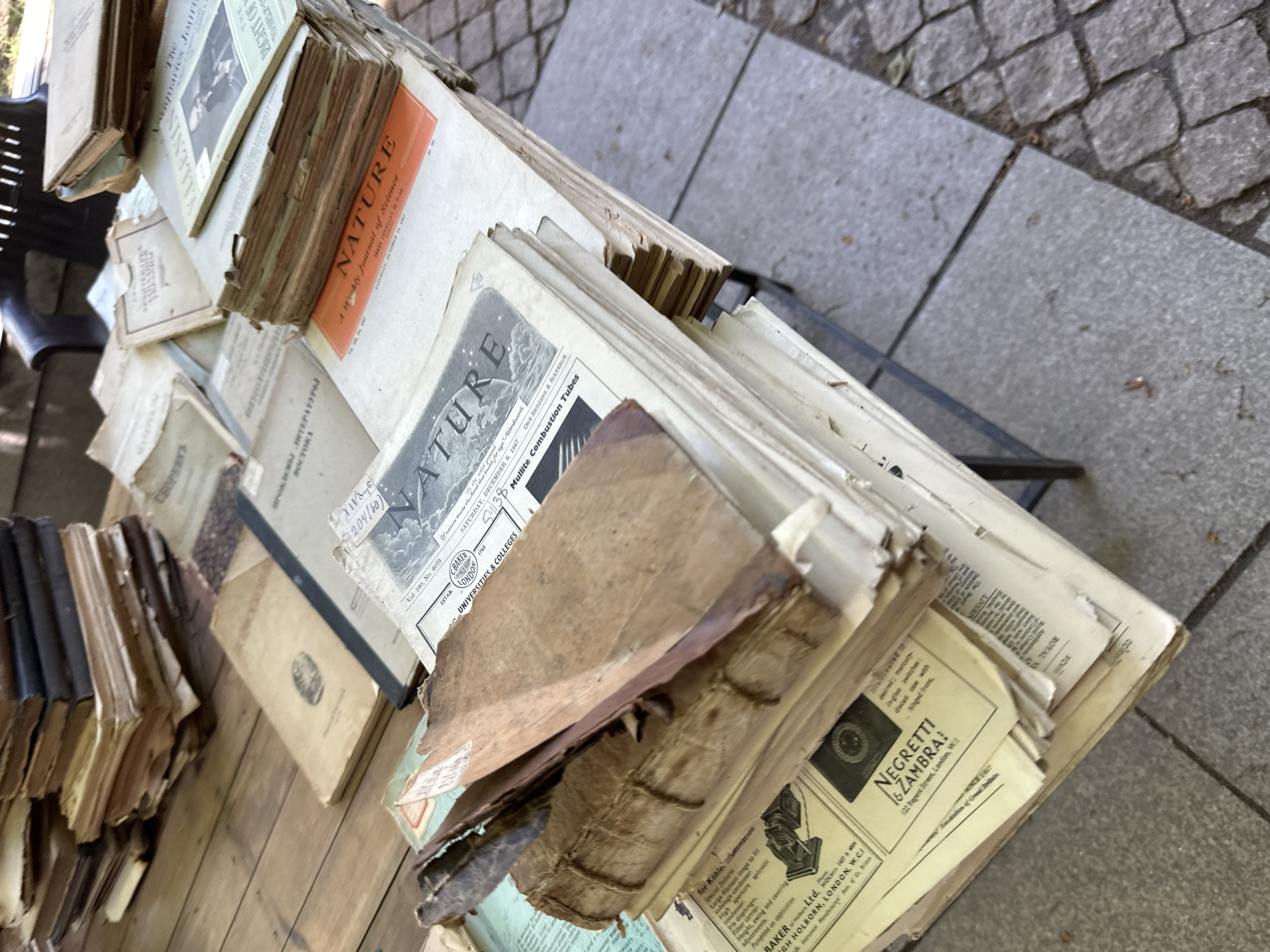 The image shows a street vendor's display of old books and magazines in Tbilisi, Georgia. Several copies of "Nature" journal are visible. The books are stacked haphazardly on a metal rack. The scene is outdoors, with a cobblestone or stone-tiled surface visible in the background. The items appear to be for sale.