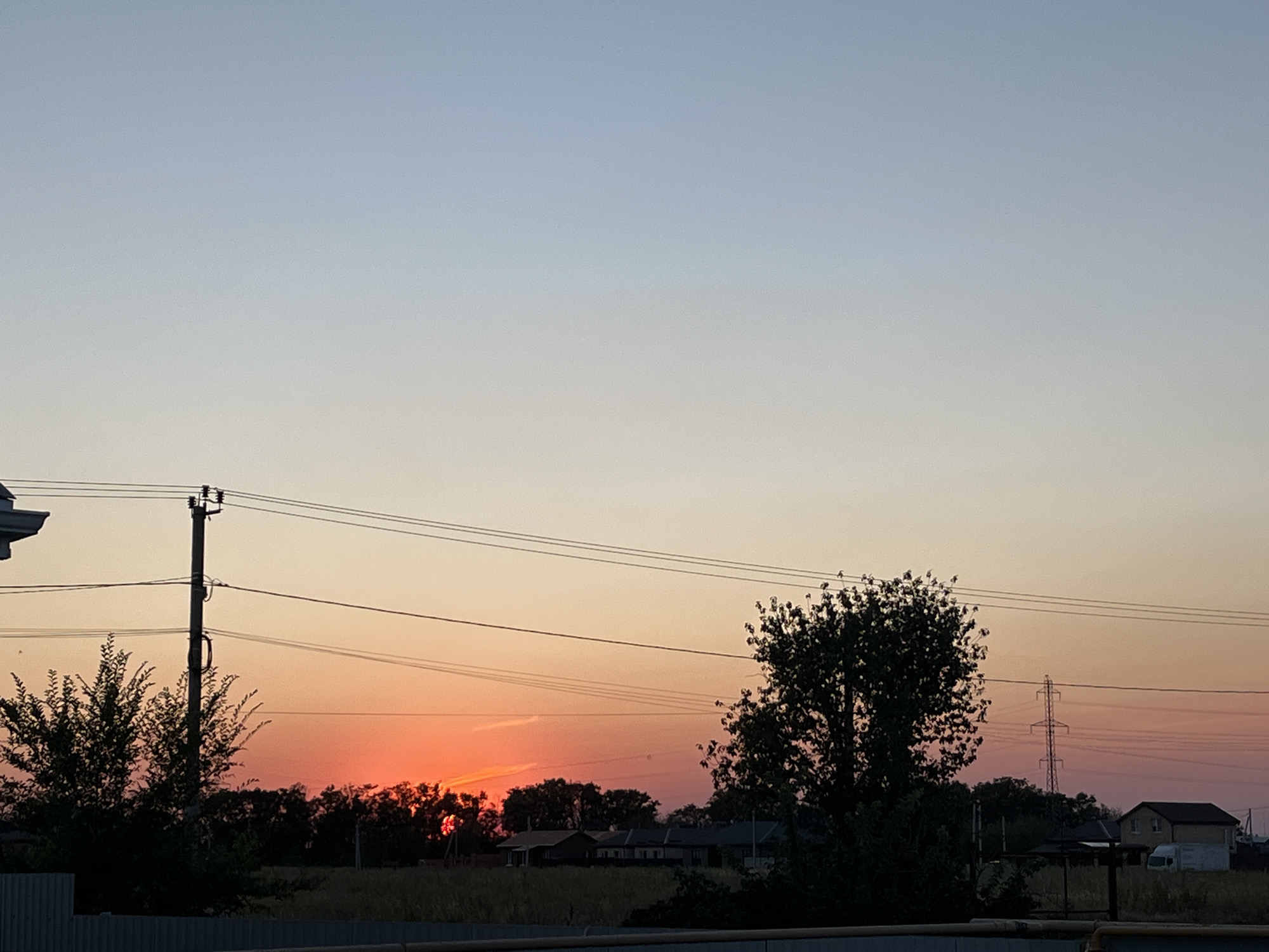 This is an outdoor, eye-level, medium shot photograph taken during sunset in Taganrog City District, Russia. The sky exhibits a gradient of colors, transitioning from a light blue at the top to shades of orange and pink near the horizon. A few power lines crisscross the middle ground of the image, supported by a tall utility pole on the left and a smaller tower on the right. Silhouetted against the vibrant sky are several trees, some appearing leafy and full, while others are more sparse. In the background, several low-rise buildings, likely residential homes, are visible. A white van is parked near one of the buildings. A fence runs along the bottom of the frame, obscuring the ground. The overall scene captures a tranquil, rural or suburban landscape at dusk.