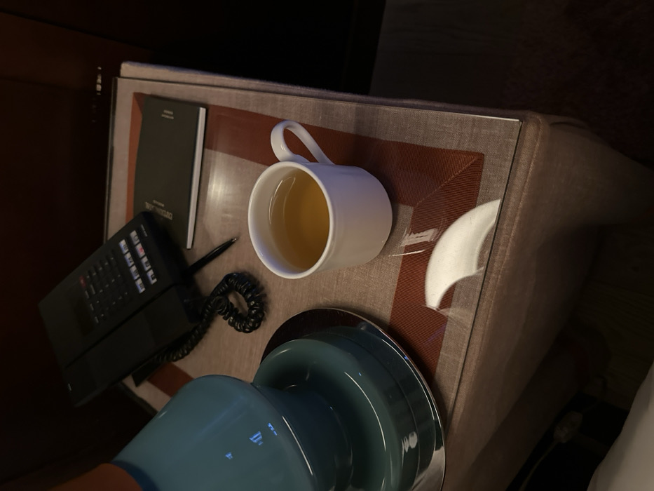 The image depicts a close-up, high-angle view of a bedside table within a dimly lit indoor environment, identified as a hotel room in Paris, France. The square table features a transparent glass surface positioned over a placemat-like cover with a beige central section and a reddish-brown patterned border. On the far left, the base of a teal or turquoise-colored lamp with a silver-toned metallic foot is visible. Towards the upper left, a black landline telephone with a coiled cord and a visible keypad rests on the table. A black pen lies horizontally between the telephone and a small, dark-colored booklet or notepad, which displays partially visible text, including "EXECUTIVE". A white ceramic mug, filled with a clear, amber-colored liquid, occupies the central foreground. The background is dark and featureless. No individuals are present, and no actions or interactions are depicted.