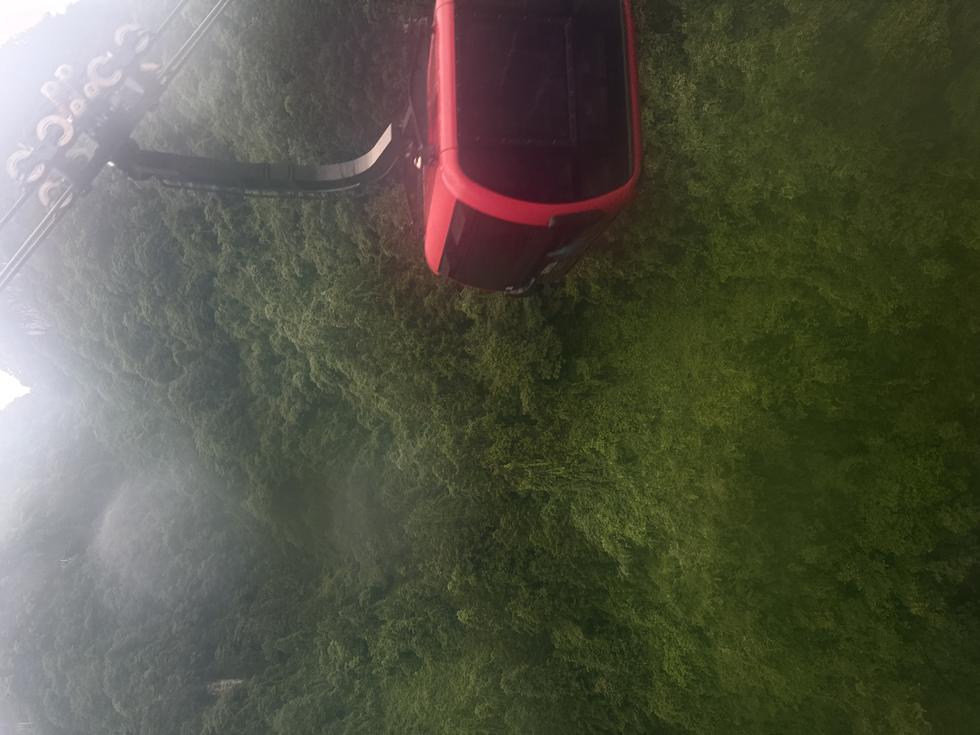 A red cable car is suspended above a lush green landscape in Yongding District, China. The aerial tramway mechanism is visible above the cable car, partially obscured by mist. The dense vegetation covers the ground below, suggesting a mountainous or hilly terrain. The image is taken from a high vantage point, likely inside another cable car.
