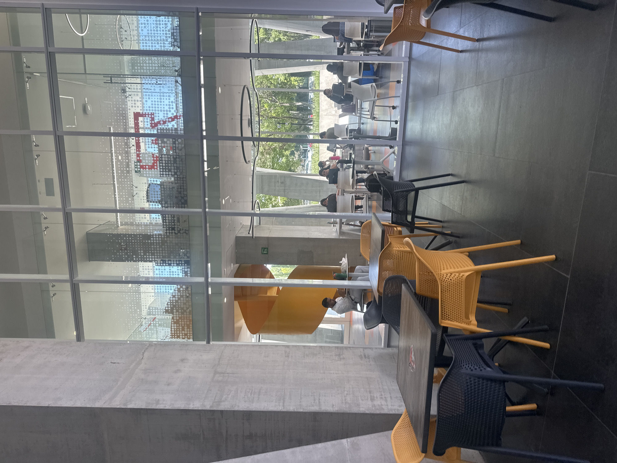 The image depicts an indoor cafeteria or dining area, likely within a larger building in San Bernardino Tlaxcalancingo, Mexico. The architecture features large glass walls, allowing natural light to illuminate the space and providing views of the exterior. The interior is furnished with tables and chairs, some of which are occupied by people.
The furniture includes black and yellow-orange chairs, and dark-colored tables.
Several individuals are visible through the glass walls, seated at tables and appearing to be engaged in dining or other activities. The area outside the building features greenery.
The image suggests a modern, well-lit dining space, integrated with the exterior environment.