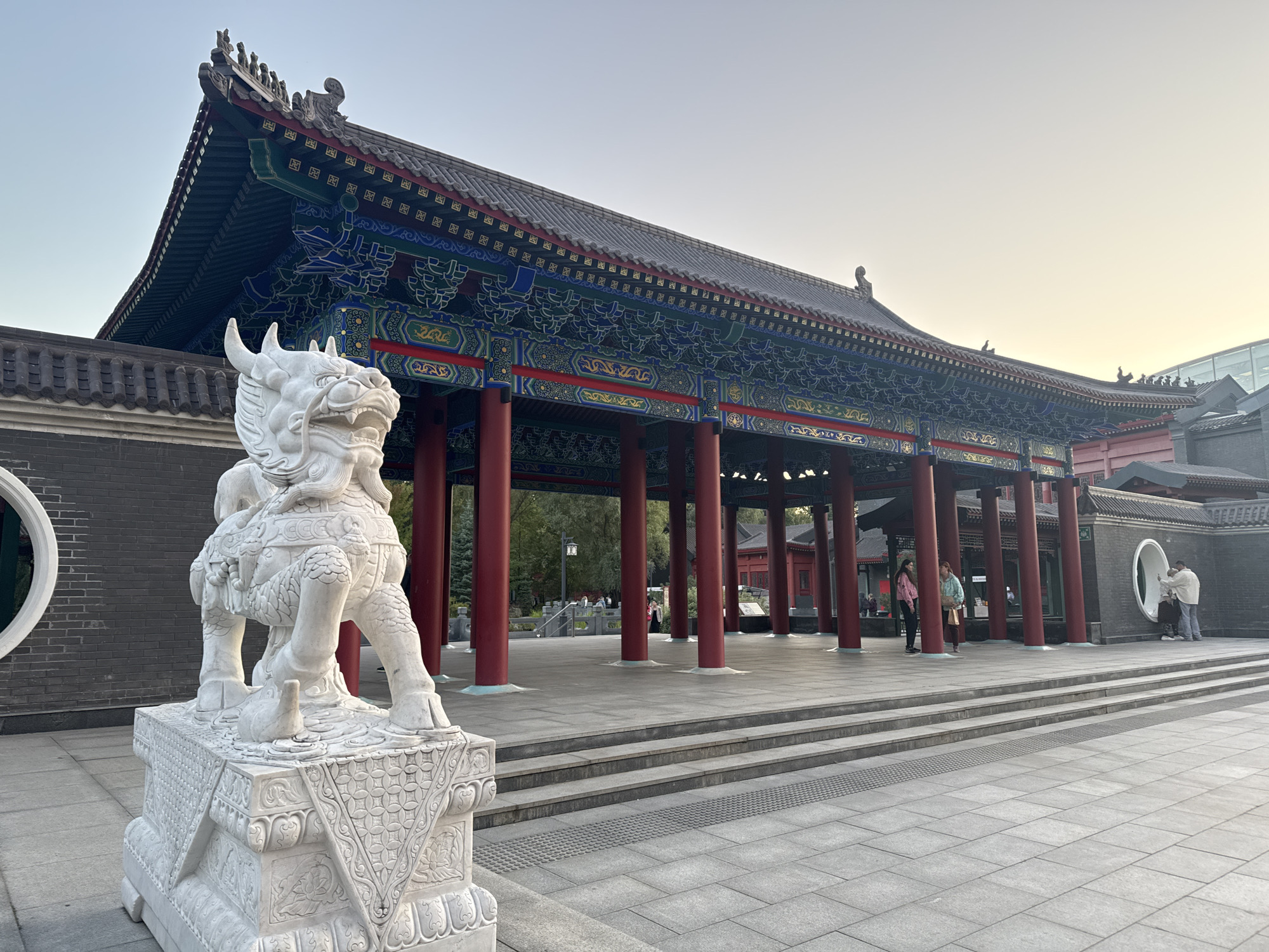 In the foreground, a white stone statue of a mythical creature, possibly a dragon or lion, stands on a carved pedestal. The statue is positioned to the left of the image, facing right. Behind it, a wide set of stone stairs leads up to a covered pavilion with a traditional Chinese architectural style. The pavilion features a dark tiled roof with ornate, colorful decorations beneath the eaves, supported by numerous red pillars. The pavilion is open on the sides, revealing trees and other buildings in the background. Several people are visible within and around the pavilion. To the left of the pavilion, a dark brick wall with a circular opening is present. The ground is paved with large stone tiles. The sky above is a pale, clear blue, suggesting it is daytime.