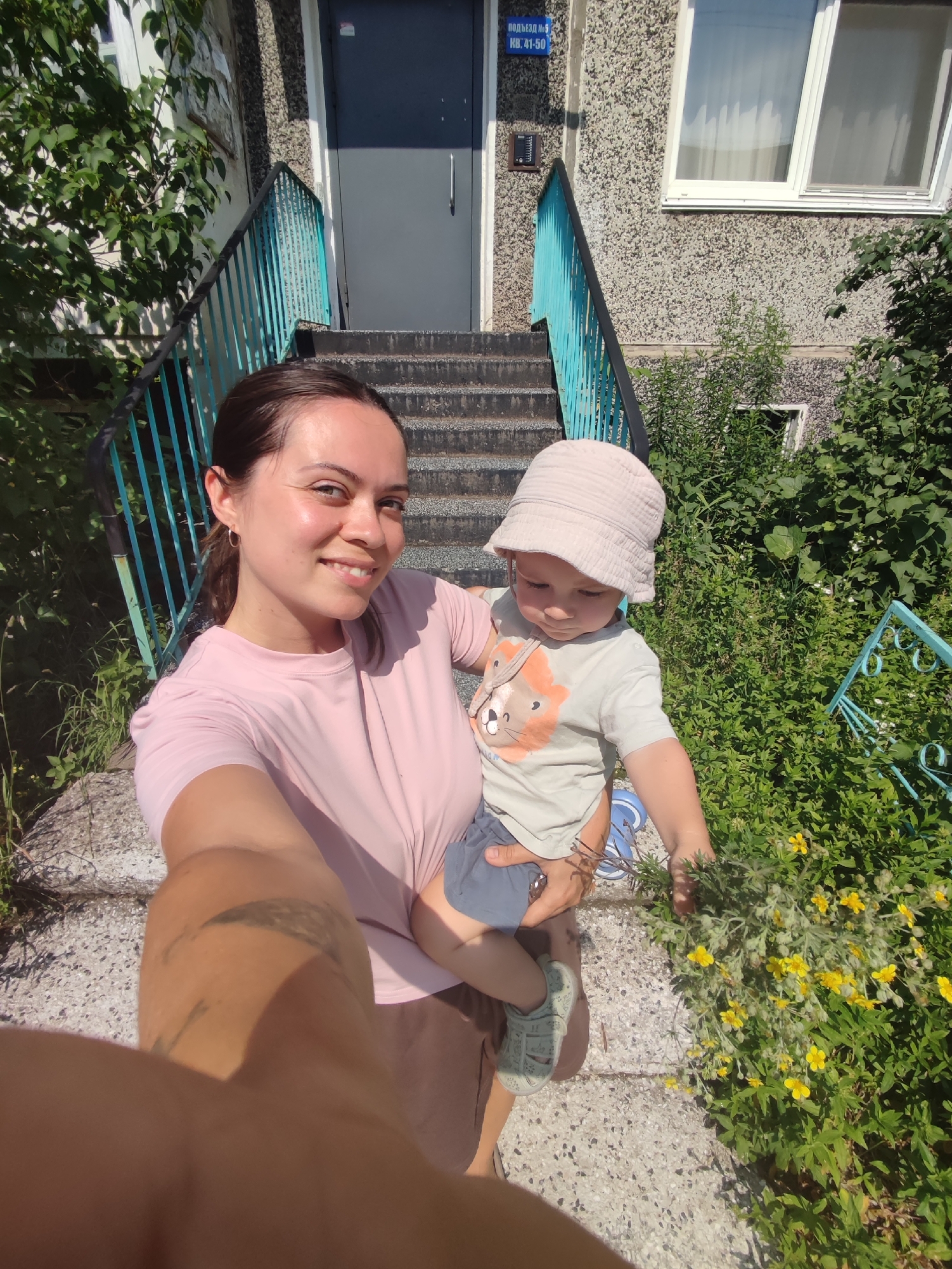 A woman and a young child are taking a selfie outdoors in front of a building. The woman, on the left, is smiling at the camera and holding the child in her arms. She has dark hair pulled back and is wearing a pink t-shirt and brown shorts. The child, on the right, is wearing a light-colored bucket hat, a light grey t-shirt with a lion graphic, and blue shorts. The child is looking down and holding a bunch of yellow flowers.
The background shows a concrete staircase with turquoise railings leading up to a building entrance. The building has a textured grey facade and a blue door. A sign above the door reads "ПОДЪЕЗД № 5" and "КВ: 41-50". This indicates the entrance to apartment building number 5, apartments 41-50, likely in Russia. Lush green foliage and small yellow flowers are growing around the base of the stairs and the building. The scene appears to be in the city of Chaikovsky, Russia, based on the sign.
