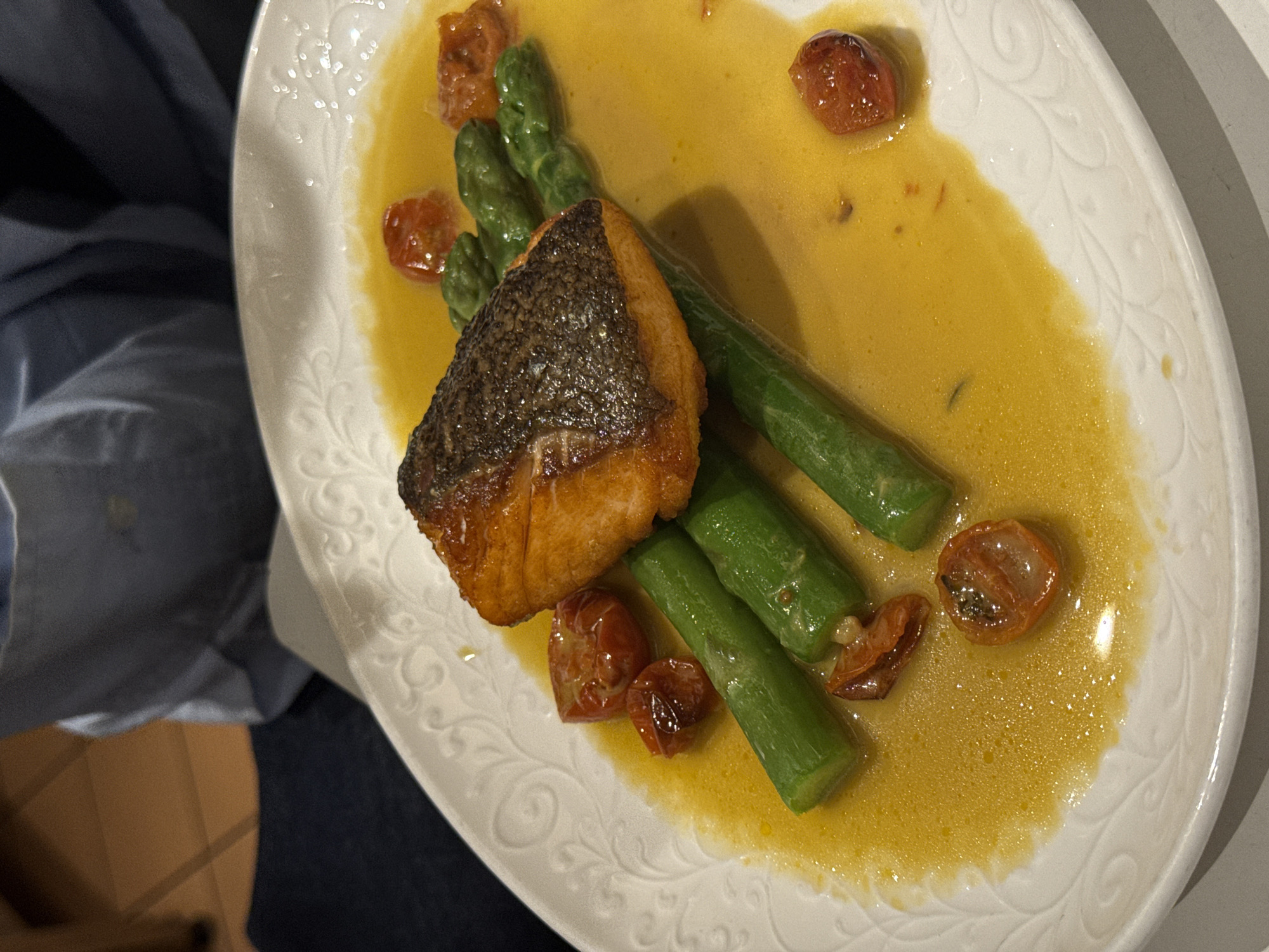 A close-up shot shows a white, decorative plate containing a piece of cooked fish with crispy skin, several spears of asparagus, and halved roasted cherry tomatoes, all covered in a yellow sauce. The plate has a raised, patterned edge. To the left of the plate, a person is partially visible wearing a blue shirt and what appears to be dark denim jeans. A wooden surface is also visible on the left. There is no indication of this being in Belgrade, Serbia.