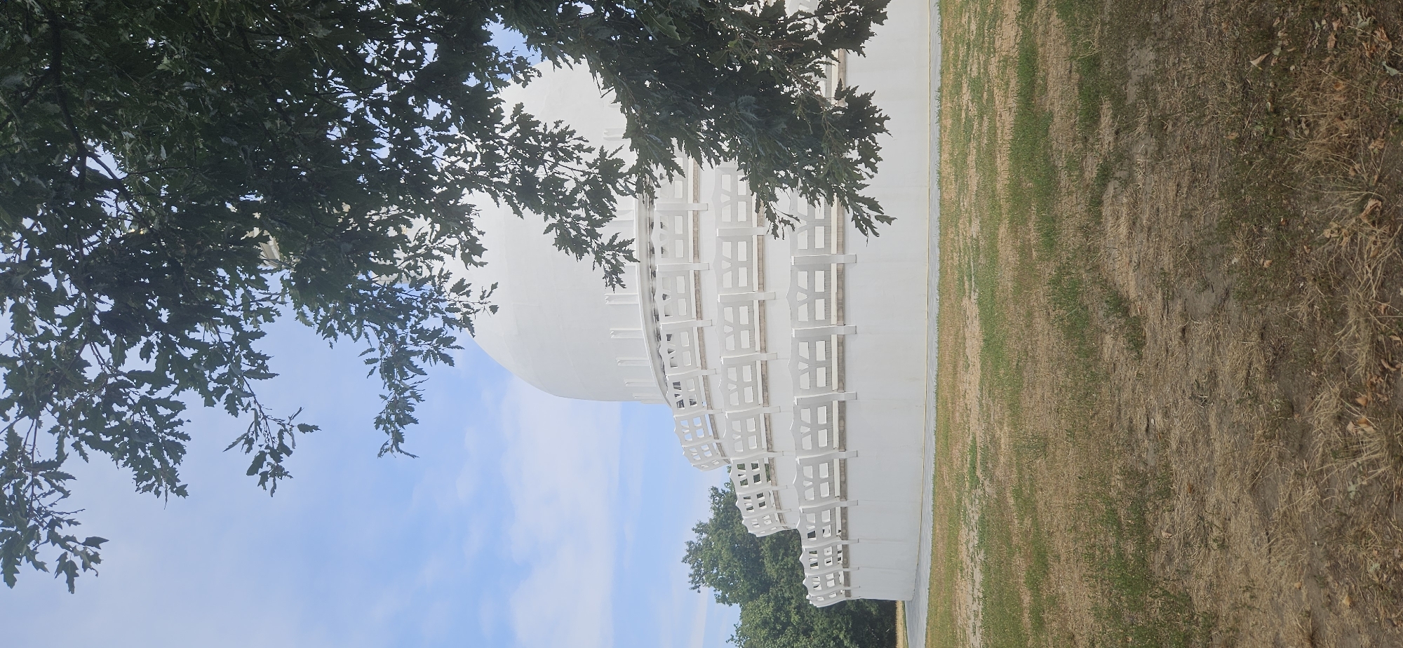 The image shows the Peace Pagoda in Zalaszántó, Hungary, a Buddhist stupa situated in a grassy, park-like area with trees. The pagoda is predominantly white, with a large dome-like structure and a tiered, white balustrade encircling its base. Several layers of the balustrade are visible, each with a repeating decorative pattern. In the foreground, dry grass and some patches of green vegetation cover the ground. Overhanging branches with green leaves from a tree partially obscure the top portion of the pagoda and the sky. The sky is a pale blue with scattered white clouds. The overall scene depicts a serene outdoor setting.