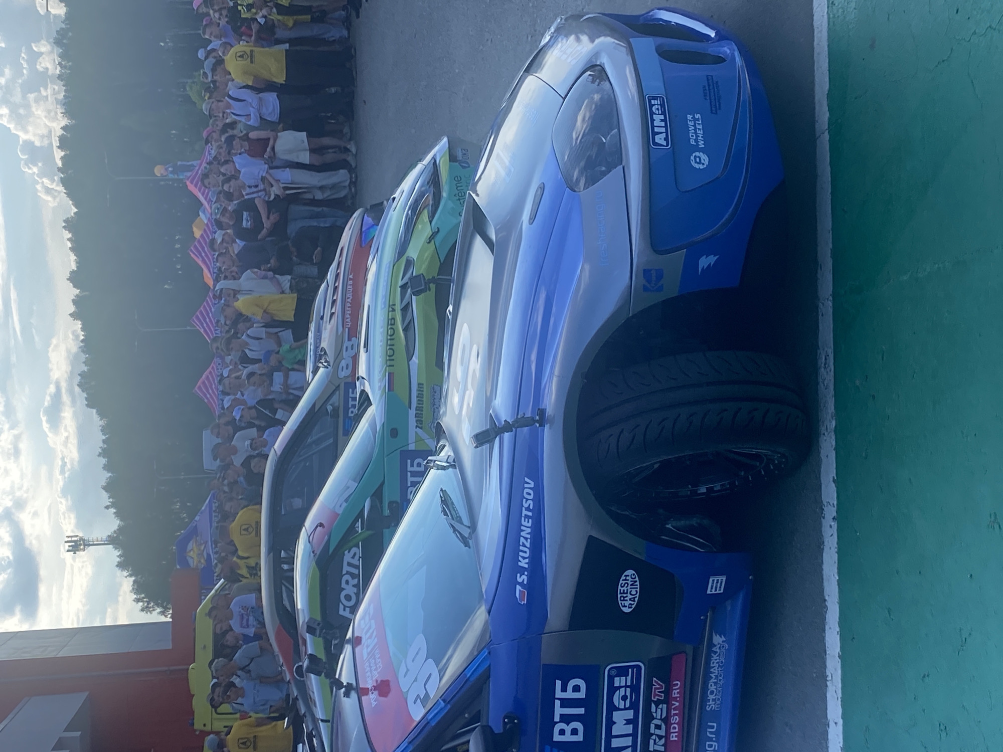 This image depicts an outdoor event with a crowd of people gathered behind a barrier, observing several race cars. Two cars are prominently featured in the foreground, with a third car visible behind them. The race cars are highly decorated with sponsor logos and racing numbers. The blue car in the closest foreground has the number "36" visible on its windshield and displays logos such as "ВТБ," "AIMOL," and "RDSTV.RU." The car behind it is green and has logos like "FORTIS" and "RTE." The background shows trees and a cloudy sky. A crowd of spectators, dressed casually, are standing and watching the scene. The event appears to be a race or car show, possibly taking place in a publicly accessible area. The text "Берёзовка, Russia" is provided as a location, but there are no specific visual cues within the image that definitively identify it as this location. The overall atmosphere suggests a daytime event with good weather.