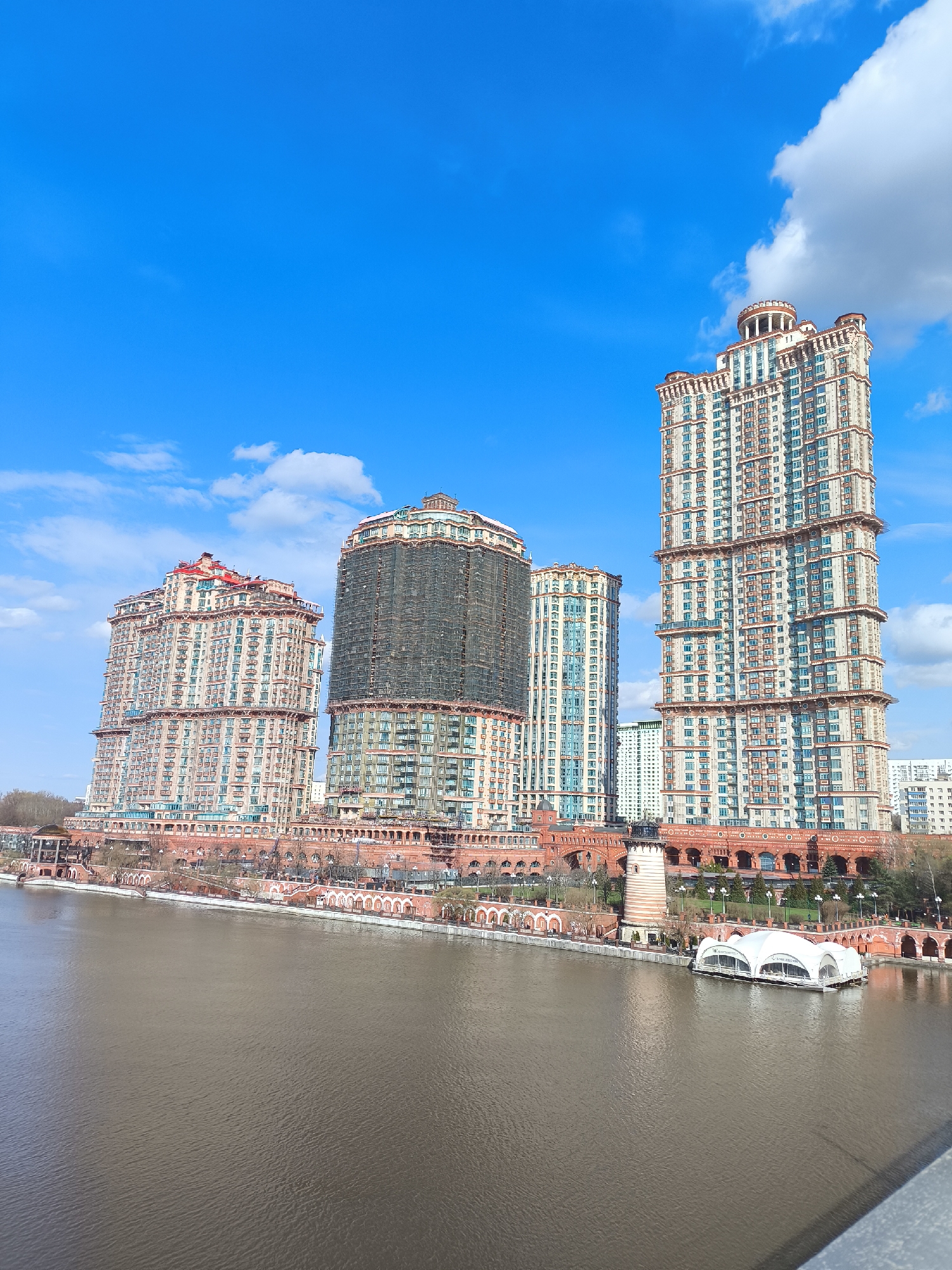 The image showcases a series of tall, ornate buildings situated along the waterfront, likely in Khimki, Russia. The buildings feature a mix of architectural styles and materials, with a prominent red brick base and intricate detailing on the facades. The building in the center appears to be under construction or renovation, as it is covered in scaffolding.
The foreground is dominated by a body of water, possibly a river or lake, with a grayish-brown hue. The sky above is bright blue with scattered white clouds. The waterfront area includes arched walkways, green spaces, and what appears to be a floating pavilion or structure.
