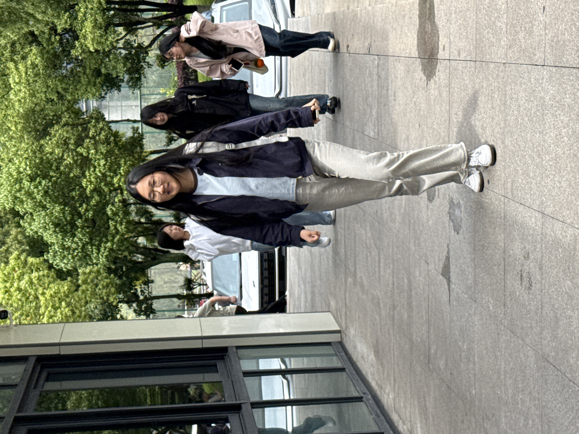 The image depicts four young individuals, likely women, standing on a paved sidewalk in an urban outdoor setting. The primary subject, a young woman with long dark hair and glasses, stands in the foreground. She is wearing a light blue t-shirt, a dark blue open jacket, and light beige or off-white loose-fitting pants, complemented by white sneakers. She is looking towards the viewer with a slight smile.
Behind her, to her left, another young woman with long dark hair is seen wearing a black jacket and dark jeans. Further back and to the right, a third young woman in a light pink jacket and dark jeans is visible, holding an orange-colored object, possibly a fruit. A fourth person, partially obscured in the background, wears a white shirt and light pants.
The background features a modern building with dark, reflective windows on the left side of the frame. On the right and in the far distance, lush green trees indicate a park-like area or tree-lined street. Several white vehicles, including what appears to be a van or SUV, are present behind the individuals. The ground is a light-colored, textured concrete or paved surface.
The scene suggests a casual daytime activity, possibly a walk or a moment of rest. The bright lighting indicates it is daytime, and the weather appears clear and pleasant. There is no visible text in the image. The overall mood is relaxed, with the prominent subject displaying a friendly expression. The location context is Jiangjin, China.
