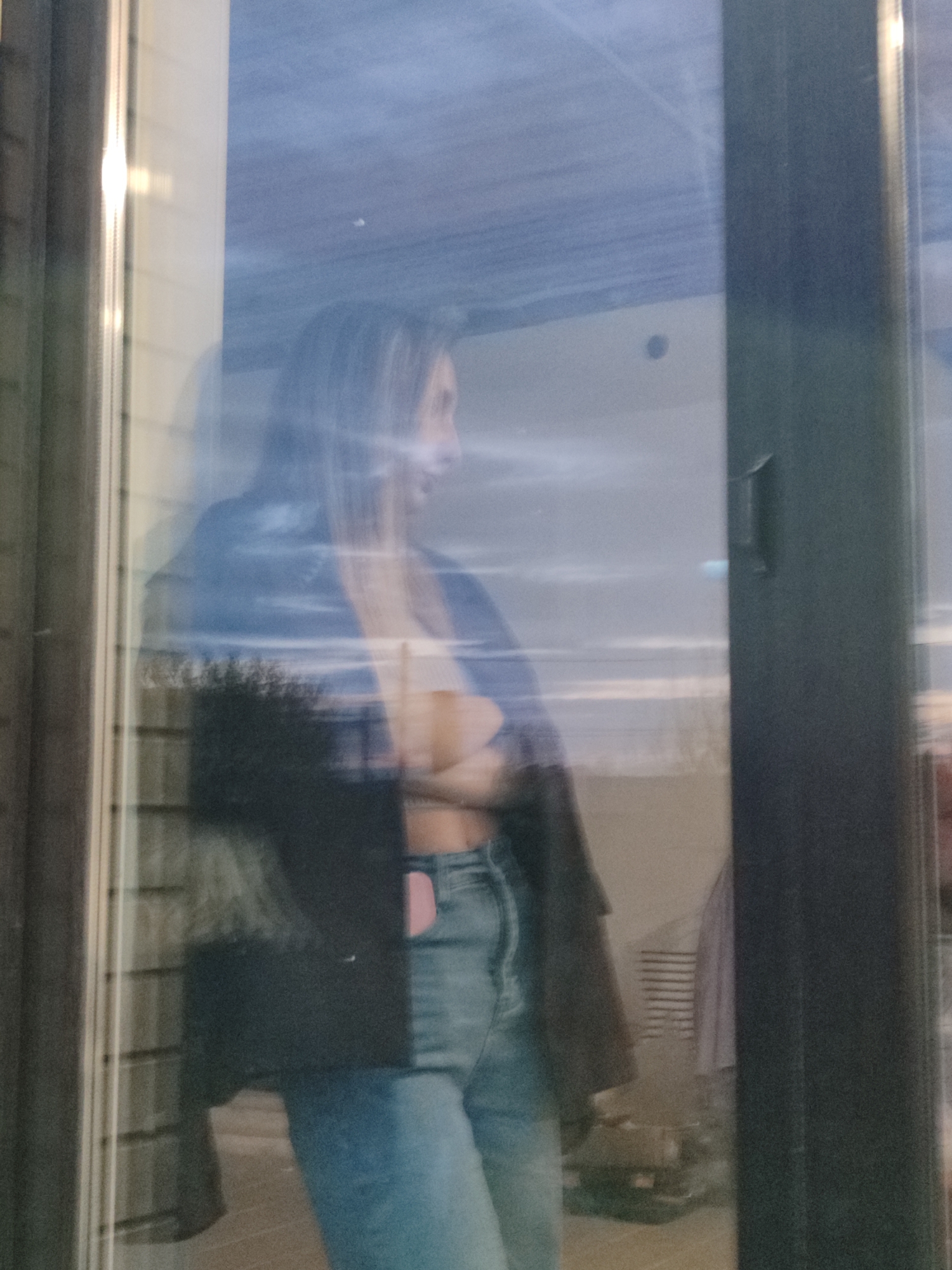 A woman is reflected in a window, seen from the outside. She is wearing a black jacket over a light-colored top and blue jeans. Her arms are crossed. The background of the reflection suggests an indoor setting with furniture and possibly some storage items. The window has a dark frame. The exterior lighting indicates it is either dawn or dusk, with a hazy sky visible. There is no specific indicator of the location being Chaikovsky, Russia.