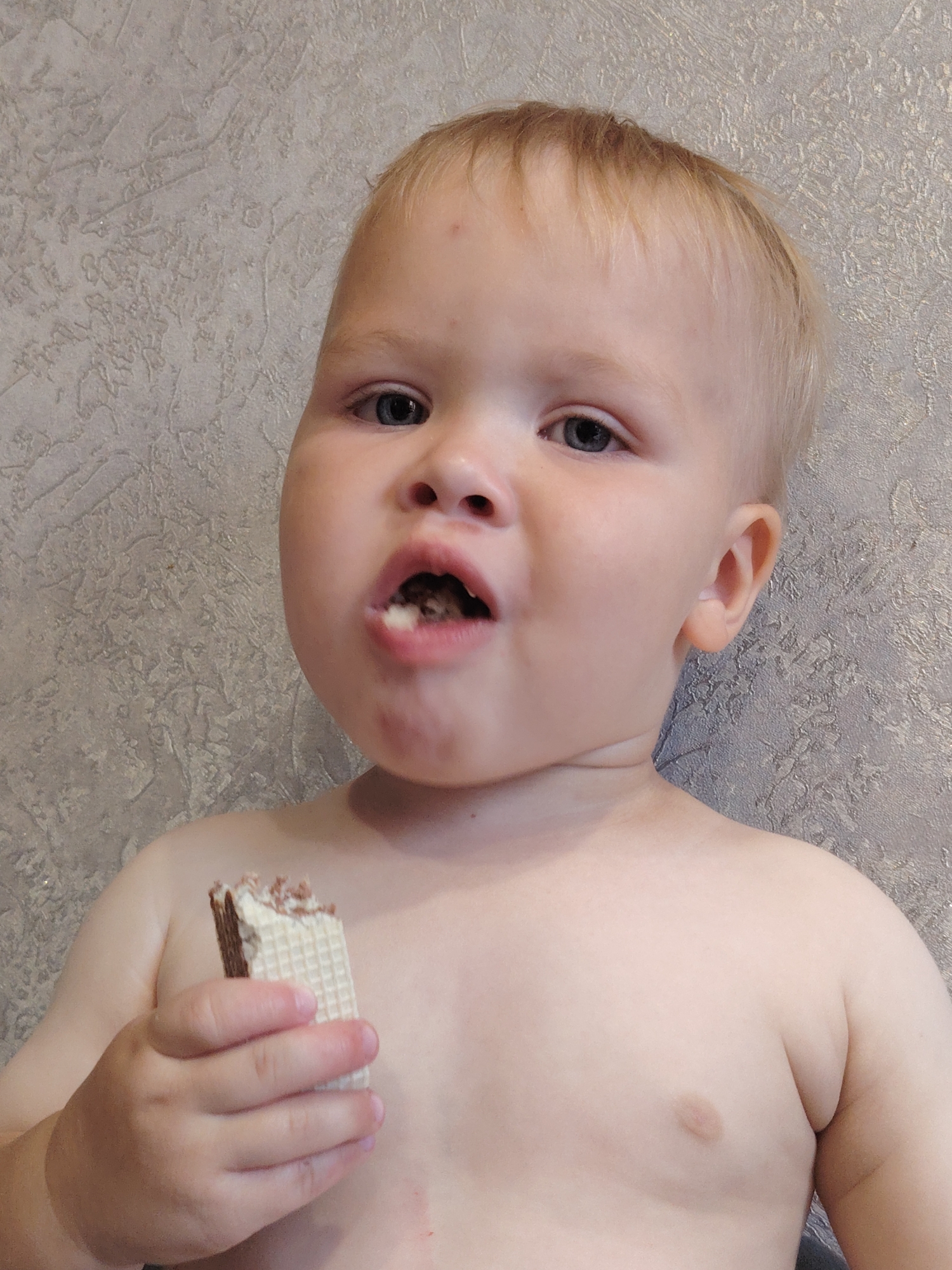 A close-up, chest-up shot shows a fair-skinned toddler with blonde hair and blue eyes, looking directly at the camera. The child's mouth is open, mid-chew, with a piece of a wafer snack in it. The toddler is holding another piece of the same wafer snack in their left hand, near their chest. The background is a textured, light-colored wall. The child is not wearing a shirt, revealing their shoulders and chest. There are no visible text elements in the image. The lighting suggests it could be daytime indoors.