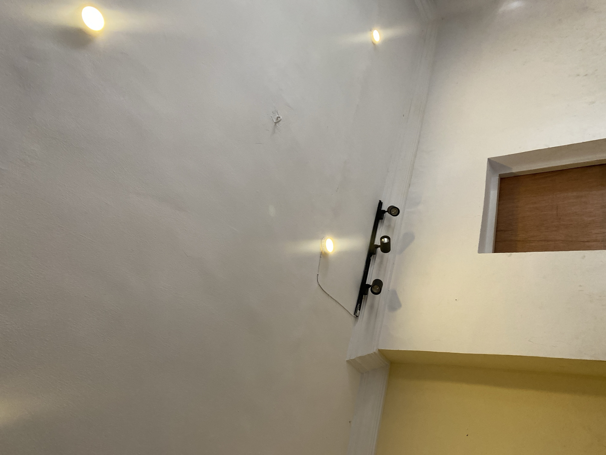 This image presents an indoor architectural scene, likely a hallway or stairwell, viewed from a low-angle perspective looking upwards. The space is predominantly characterized by textured white walls, with a section of a muted yellow wall appearing in the lower right.
Several artificial light sources illuminate the area. Two circular, warm-white light fixtures are directly mounted on the white wall: one in the top left corner and another positioned mid-right. A black track lighting system is installed vertically on the upper right side of the white wall, featuring at least two black spotlight heads that cast directed light downwards. A thin white wire connects the mid-right circular light fixture to this track system.
In the upper right quadrant of the image, there is a rectangular opening, framed with light-colored trim, revealing a dark wooden panel or shutter. Below this opening and extending across the lower portion of the right side, the wall transitions to a yellowish hue, indicating a different architectural element or a lower section of the wall.
The white walls display a slightly uneven, textured finish. A small, light-colored object, possibly a spider, is visible on the central white wall. The artificial lighting suggests an indoor setting where natural light is either absent or insufficient, implying it could be evening or a functional, interior space. There is no visible activity, people, or text in the image. The location context provided is Badagry, Nigeria.