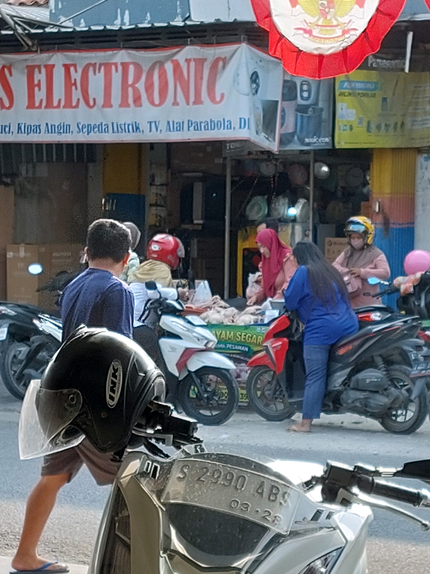 The image shows a street scene in Sumuragung, Indonesia, with a storefront advertising electronics and other goods. The storefront features a large banner with red lettering that reads "S ELECTRONIC" and below it, smaller text listing items such as "kunci, kipas angin, sepeda listrik, TV, alat parabola." Several people are visible in the scene. In the foreground, a person wearing a blue shirt and shorts stands next to a parked motorcycle, with a black helmet in the immediate foreground. Behind them, several other individuals are interacting at a stall selling what appears to be raw chicken. These individuals are dressed in casual clothing, and some are wearing headscarves or helmets. Motorbikes are parked along the street, indicating a typical urban environment in Indonesia. The background shows more shops and buildings, typical of a commercial area. The lighting suggests it is daytime, with sunlight casting shadows.