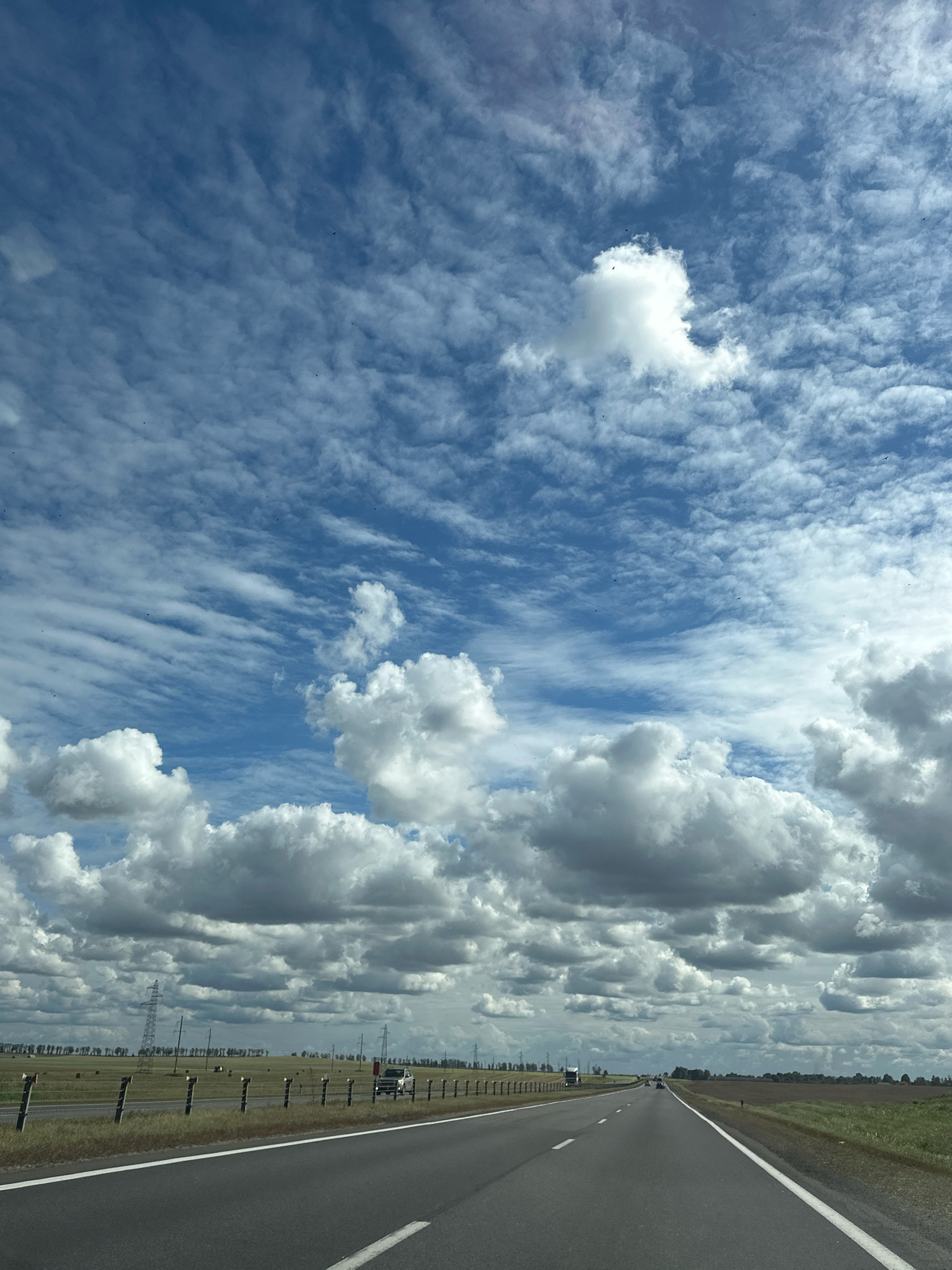 The image shows a rural landscape with a road and a dramatic sky. The road is asphalt with white lane markings and curves slightly to the right. To the left of the road, there is a grassy field with several hay bales and tall power line towers. A fence runs along the edge of the field. A silver SUV is visible on the road to the left. The sky is filled with a mix of cumulus and cirrus clouds, with patches of blue sky visible. The overall impression is of an open, natural environment, possibly in a rural area of Турынскі сельскі Савет, Belarus, given the presence of power line towers, which are common in such regions.