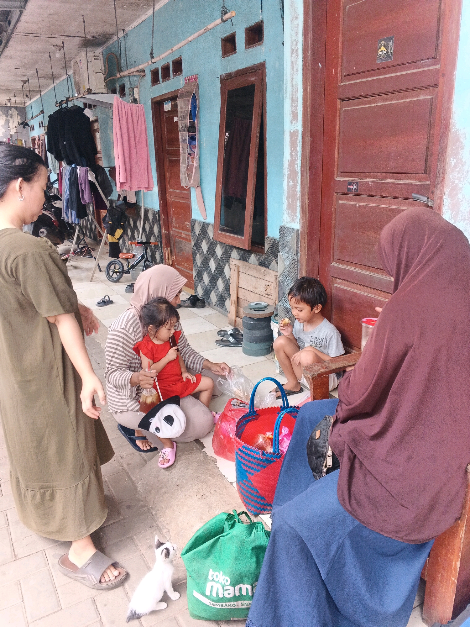 In front of a light blue building with red wooden doors and windows, a group of people are gathered on a paved area. A woman wearing a green dress stands on the left, facing a seated woman in a striped shirt and headscarf. The seated woman holds a toddler wearing a red dress, who is holding a straw with a drink. Next to them, a young boy in a grey t-shirt sits on a wooden bench, eating a snack. Another woman in a dark purple headscarf and blue skirt is seated on the right. A black and white cat is on the ground near a green shopping bag. A red and blue woven basket is filled with items. Clothes are hanging on a rack in the background, and a small bicycle is visible near a motorcycle. The location appears to be a residential area in Kadu, Indonesia.