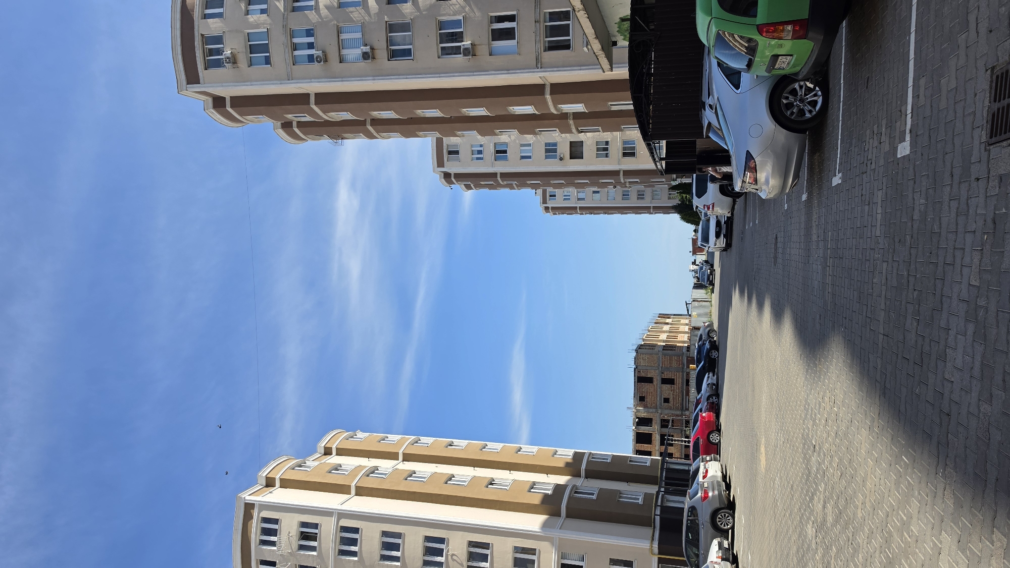 An eye-level, wide shot captures a street view with apartment buildings and a parking lot. Two tall, modern apartment buildings, one on the left and one on the right, are visible. The building on the left is tan with brown accents, and the building on the right is tan with brown trim, featuring multiple windows and air conditioning units. In the background, a partially constructed building stands, revealing brickwork and metal framing. Several cars are parked in a lot paved with gray brick pavers. The sky is a clear blue with sparse, wispy white clouds. The scene is bathed in daylight, casting shadows from the buildings onto the parking lot.