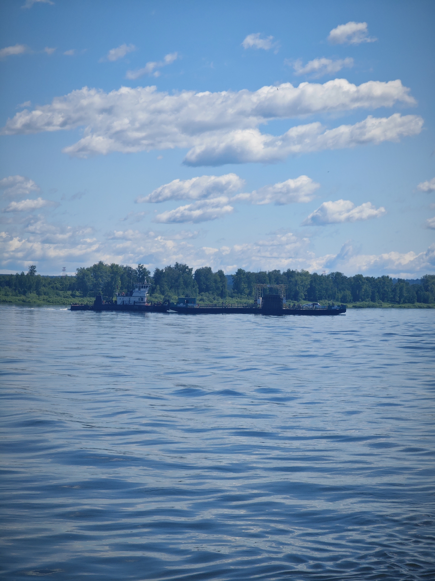 A wide, blue river with gentle ripples dominates the foreground. In the middle ground, a long, dark-colored barge is being pushed by a white and black tugboat. The tugboat has a cabin structure with windows. The barge appears to be carrying some sort of industrial equipment. A treeline of green trees forms the background, with a bright blue sky filled with scattered white clouds above. The scene suggests a sunny day in a rural or semi-rural location, possibly on the outskirts of Krutoi Log, Russia, given the industrial nature of the vessel and the natural landscape.
